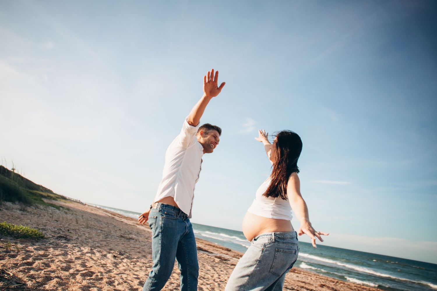 Pregnant couple dancing joyfully on a sunny beach, man raising hand, woman in crop top showing baby bump.