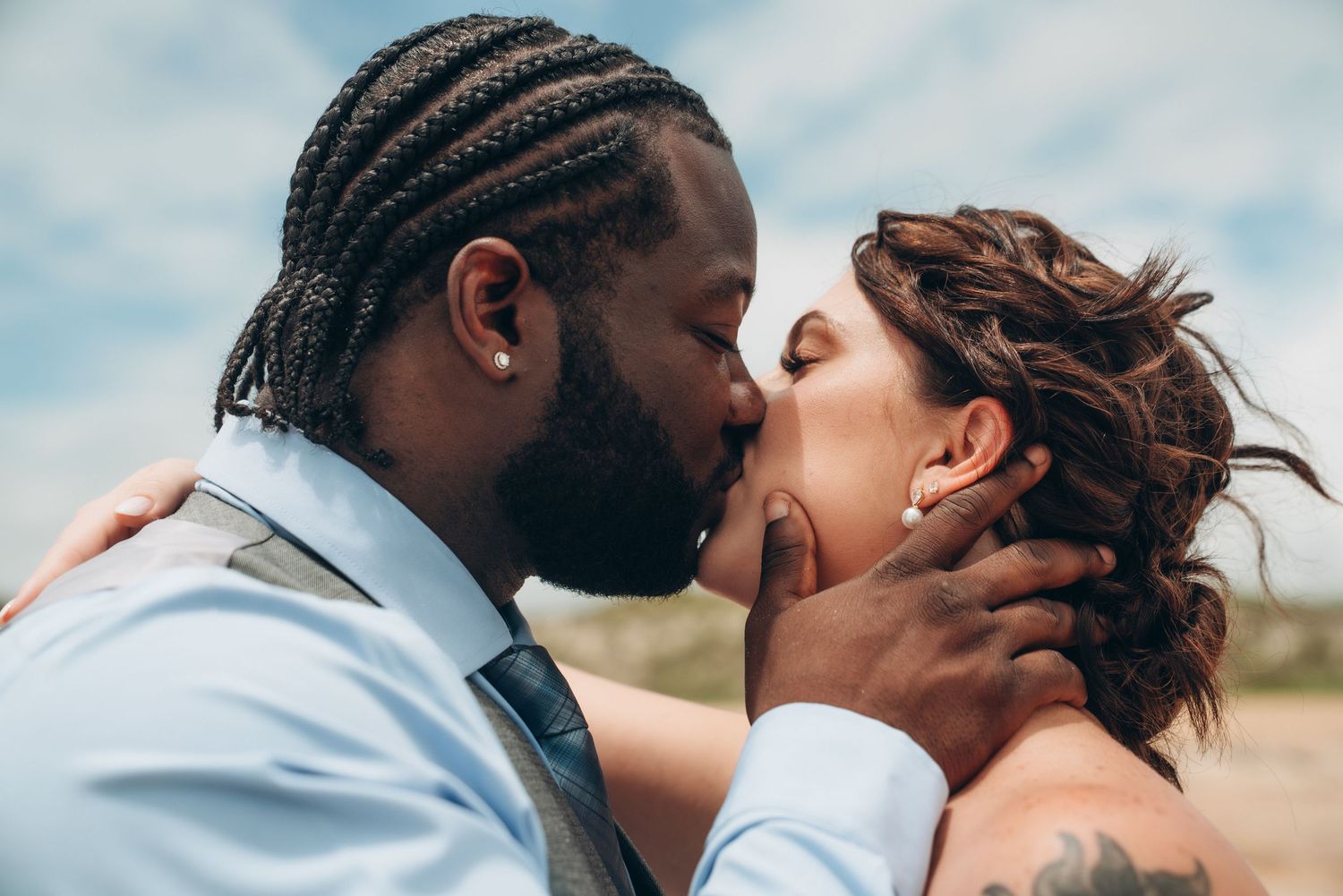 Couple sharing a romantic kiss outdoors on their wedding day, groom in blue shirt with braids, bride with updo hairstyle.