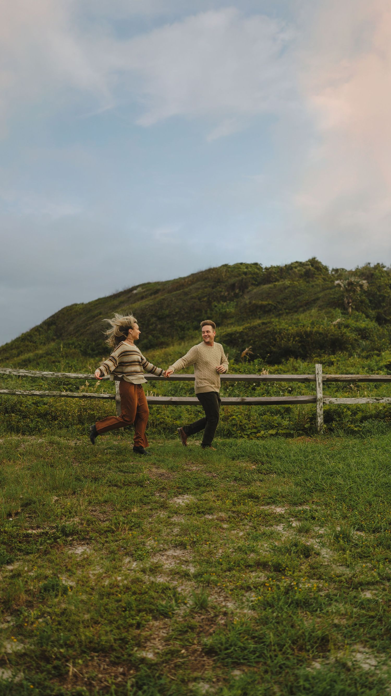A couple playfully runs along a grassy hilltop with wooden fencing and blue skies.
