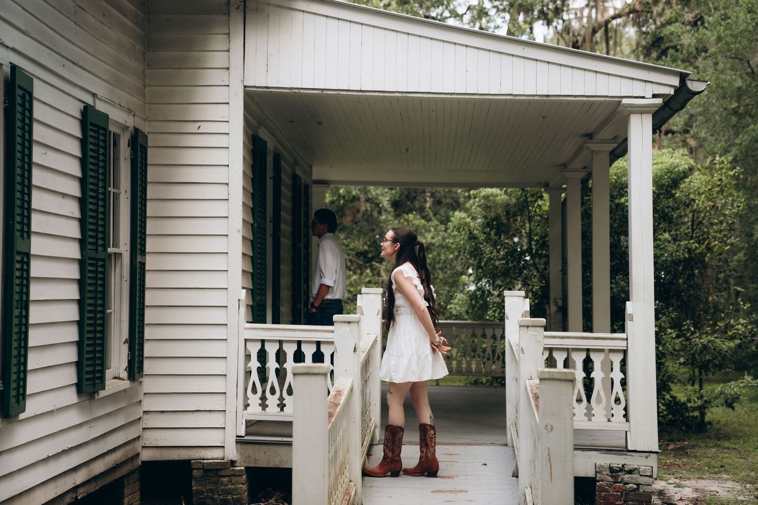 White colonial-style house with wraparound porch and green shutters in a garden setting.