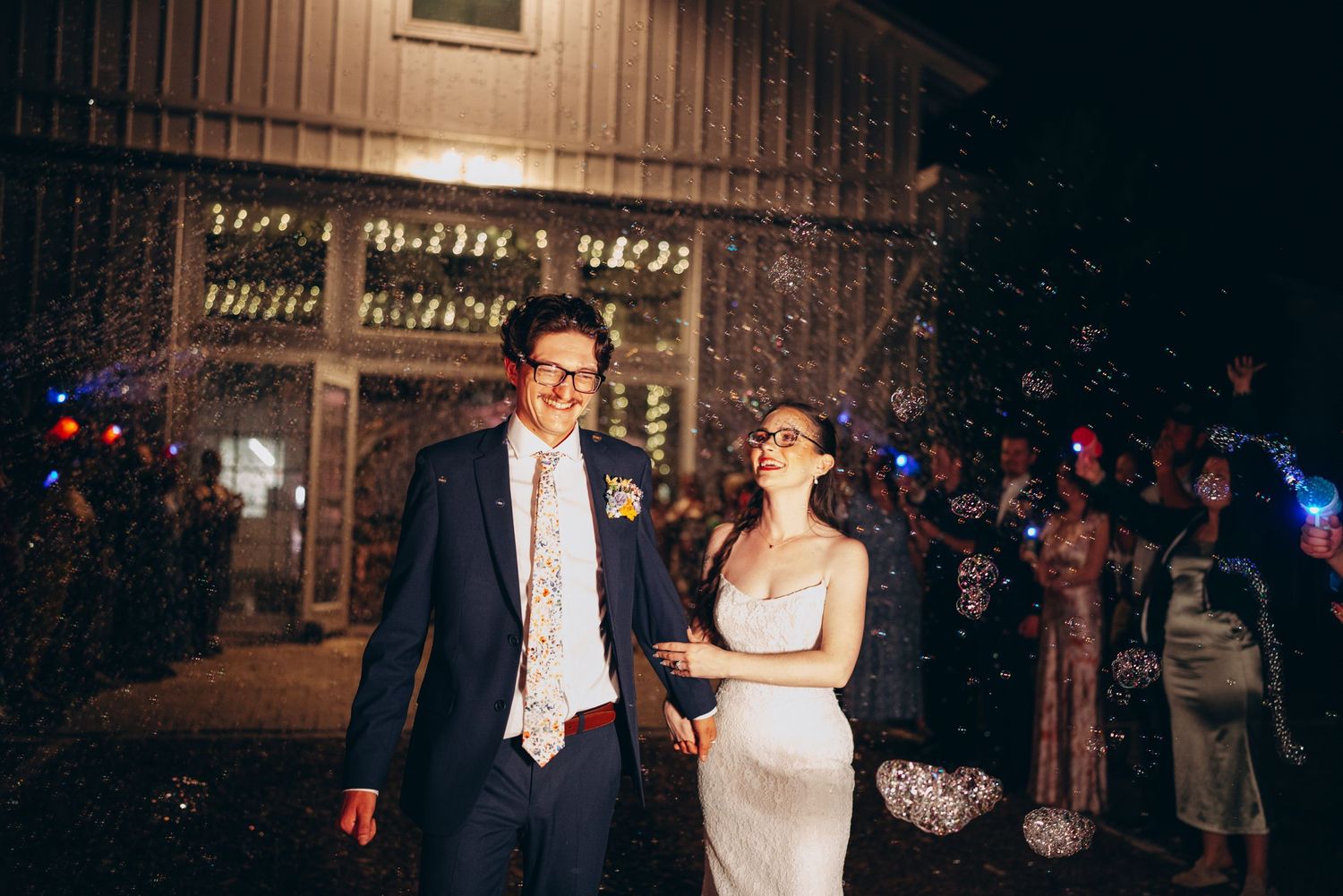 Bride and groom walk hand in hand through confetti shower at night outside rustic wedding venue.