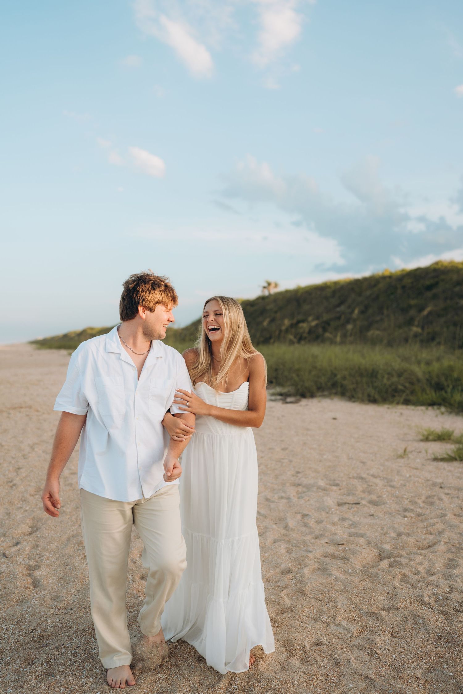A couple laughs together while walking on a sandy beach path near green hills.
