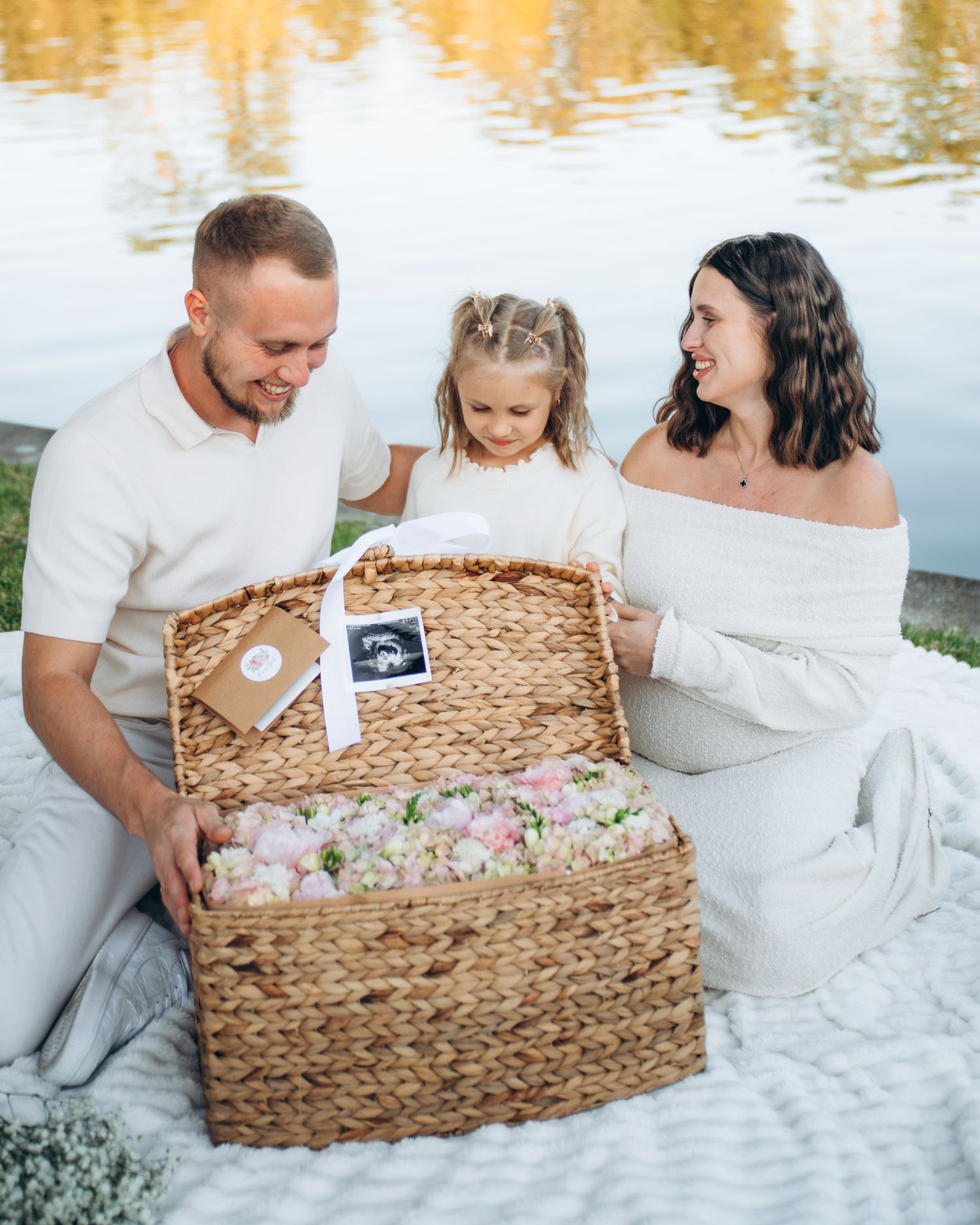 Family pregnancy announcement with ultrasound photo in a wicker basket filled with flowers by a lake.