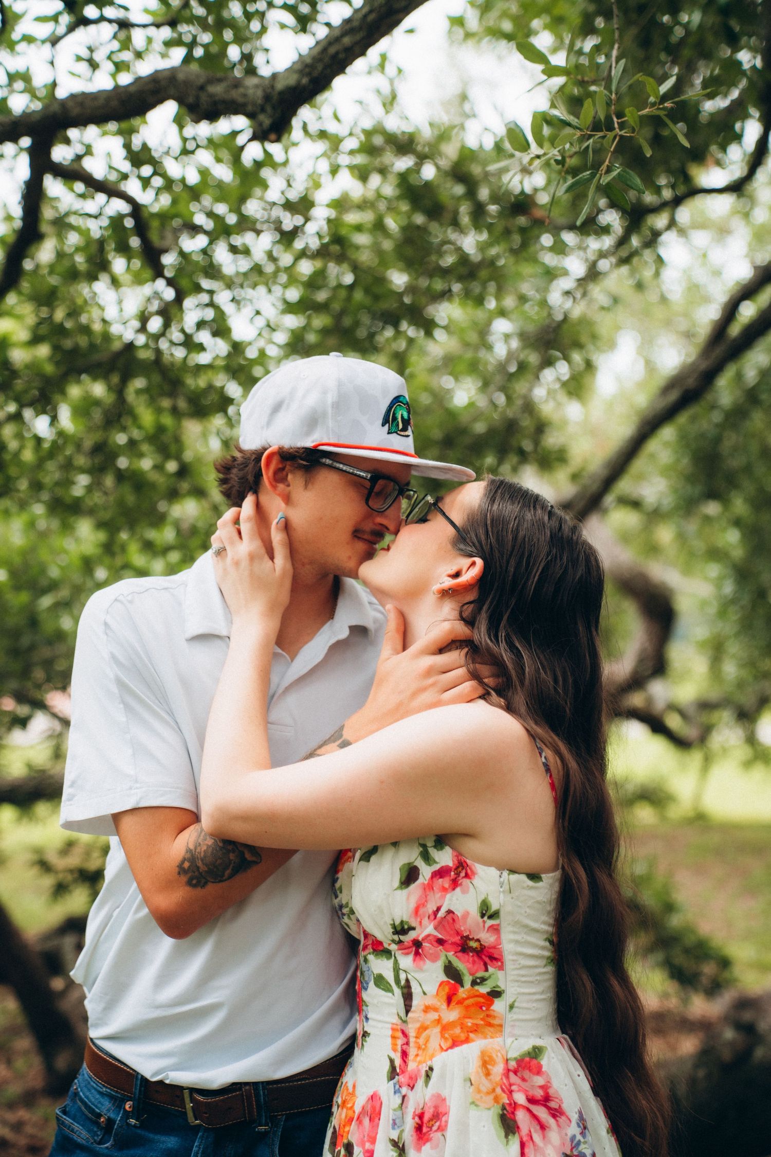 Couple sharing a romantic kiss under green trees, woman in floral dress and man in white shirt.