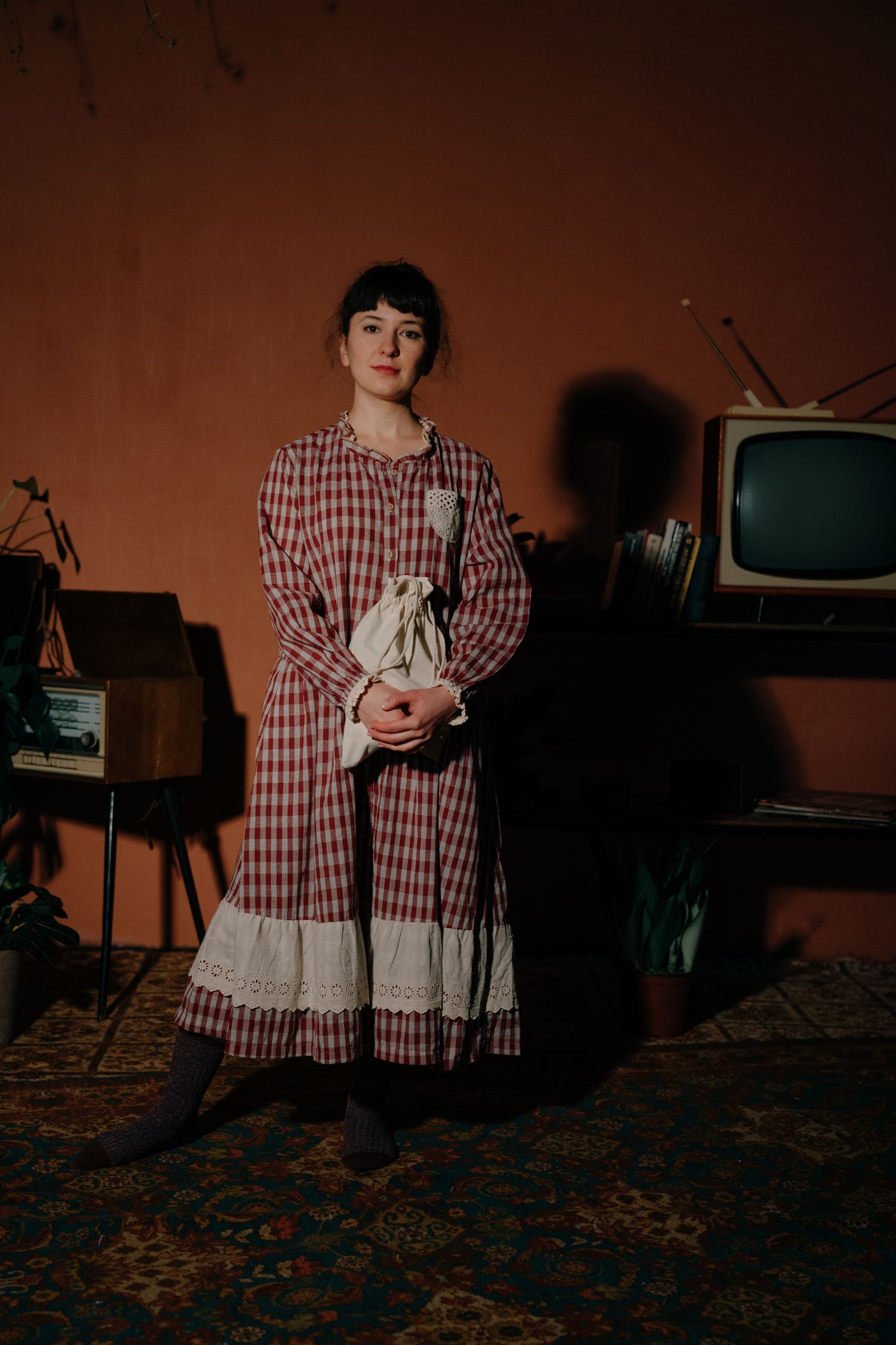 Woman in plaid prairie dress holds fabric bag, standing before vintage TV and record player on patterned rug.