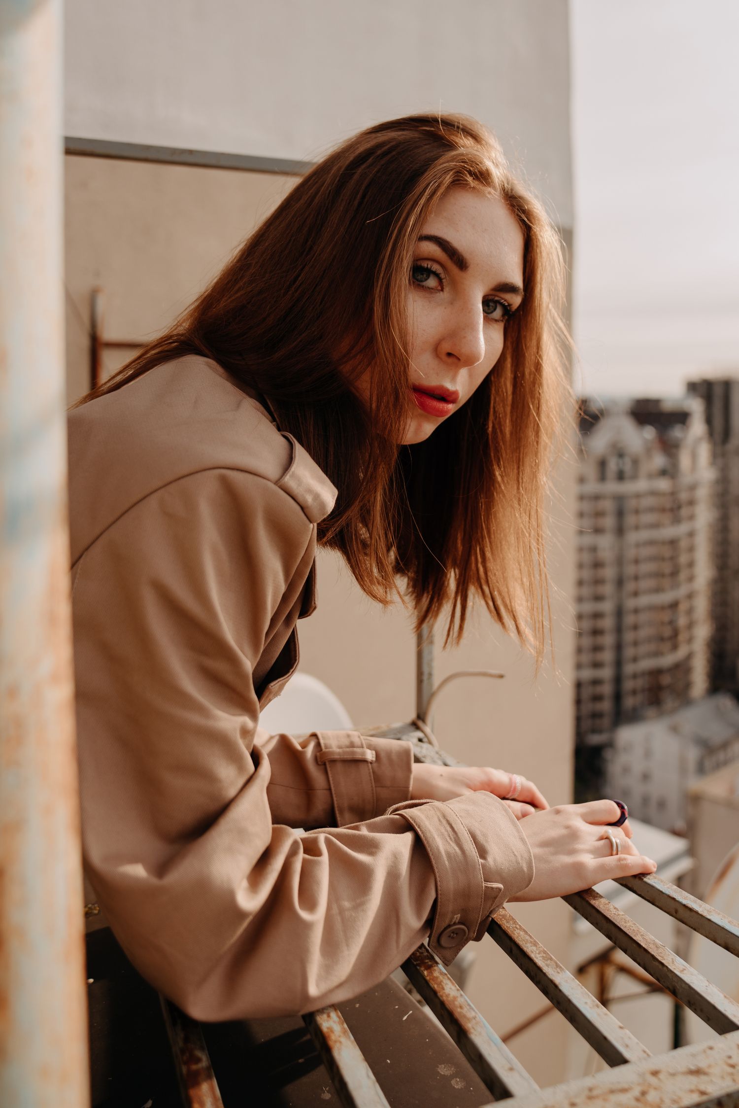 Young woman with red hair in beige coat leans on urban balcony railing, looking over shoulder at camera.