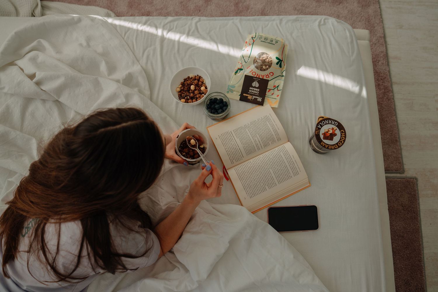 Woman reading an open book on a white bed surrounded by snacks, berries, and a smartphone in natural light.
