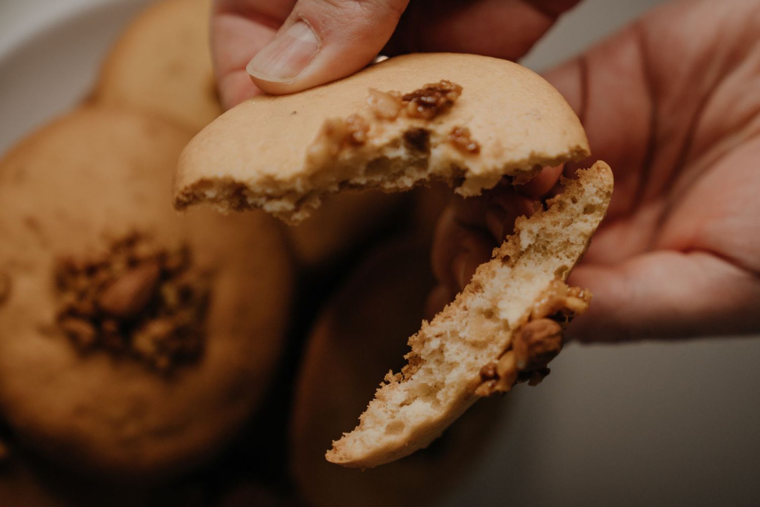 Hands breaking open a golden brown cookie revealing a chewy filling, with more cookies on a plate in the background.