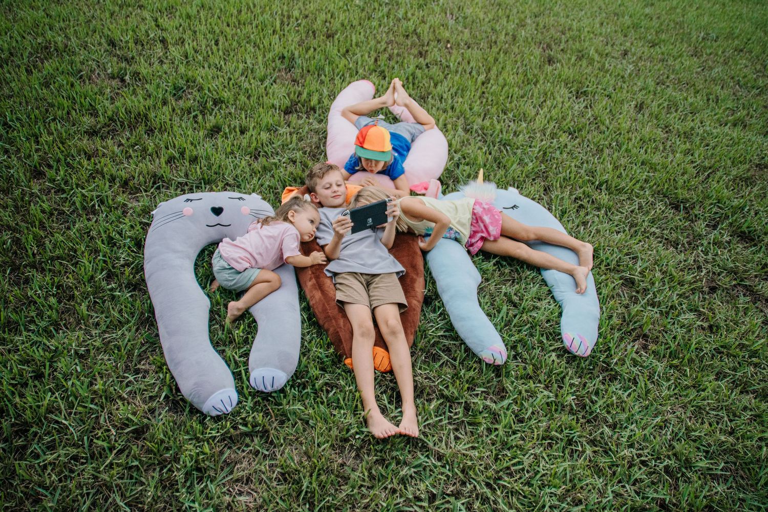 Four children lying on green grass in a star shape with colorful stuffed animal pillows outdoors.