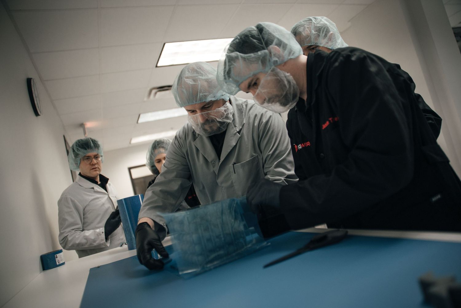 Group of researchers in hairnets and lab coats collaborate over equipment on a blue table in a cleanroom.