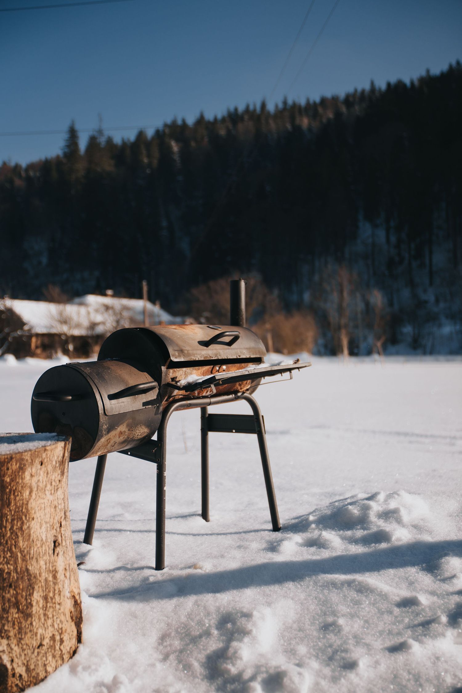 Vintage barrel BBQ smoker grill on snow with forested mountain backdrop in winter.