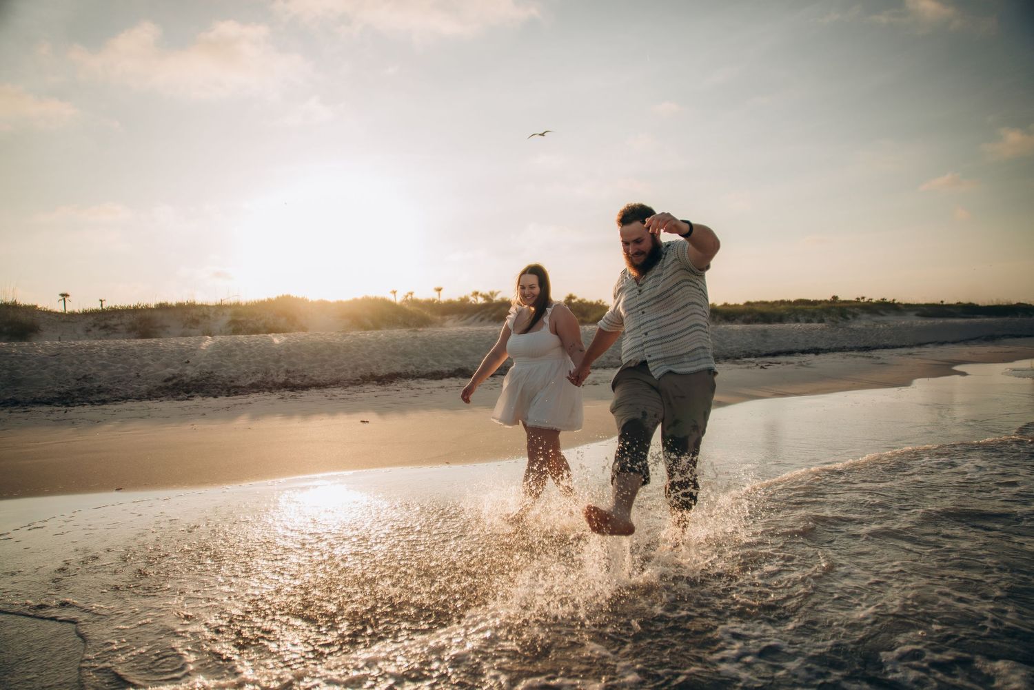 Joyful couple splashing through shallow water at sunset on beach.