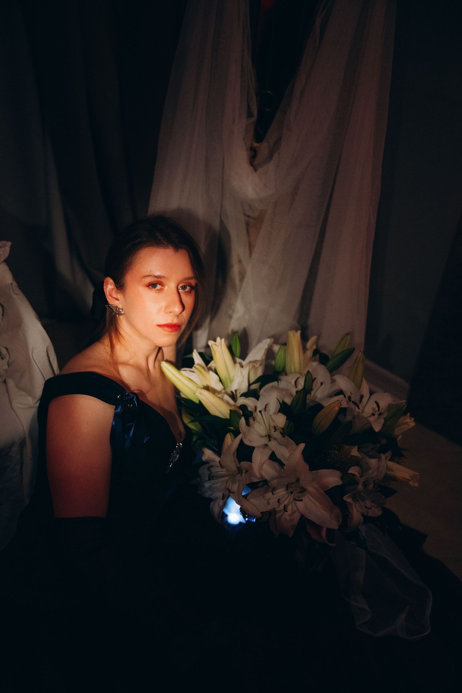 Woman in black dress holding white lilies bouquet in dramatic dark setting with flowing white fabric backdrop.