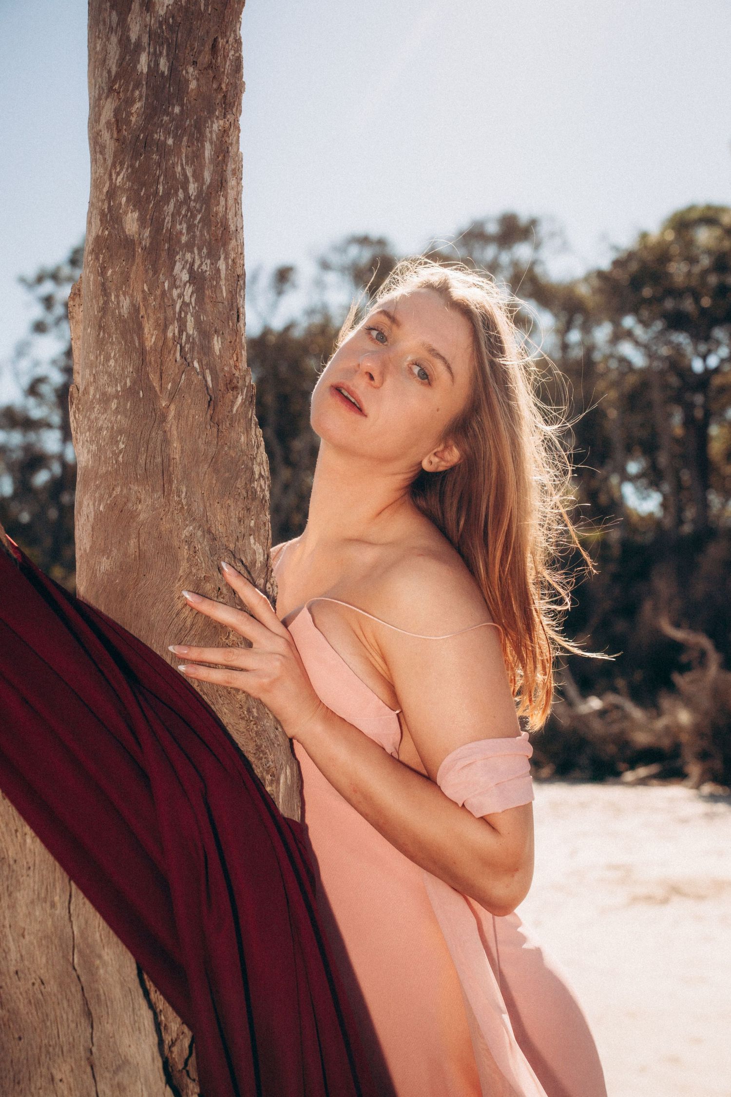A person in a peach-colored dress poses expressively against a tree trunk in golden sunlight.
