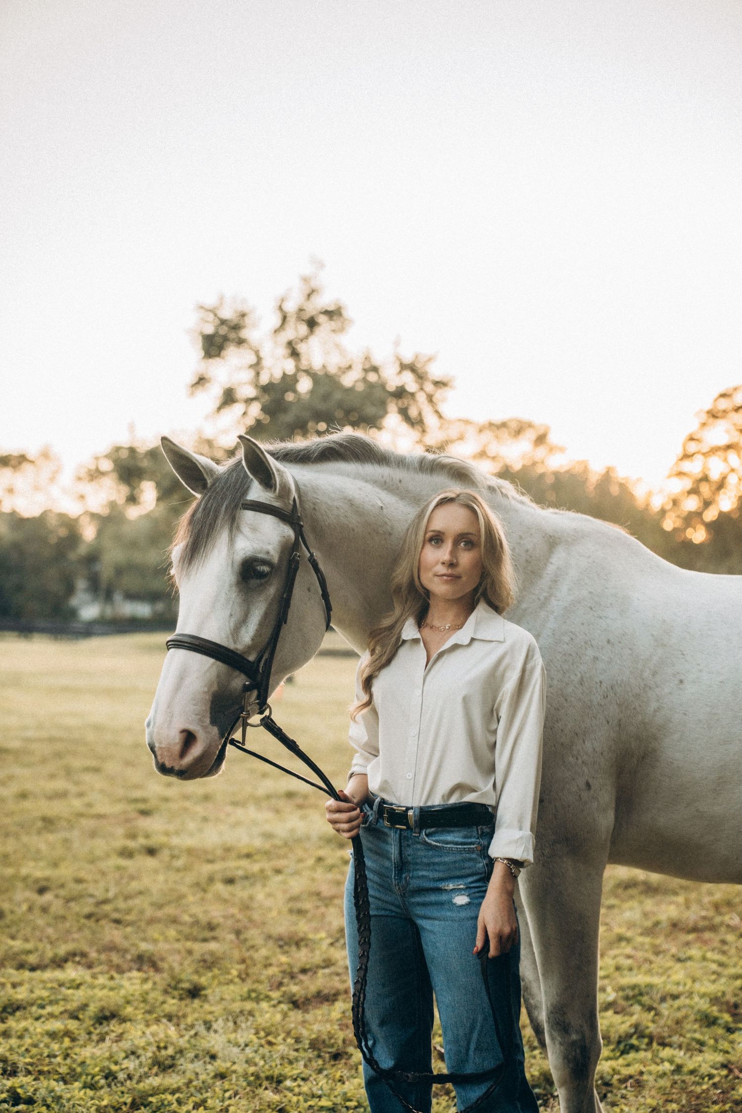 A beautiful white horse stands with its handler in a grassy field during sunset.