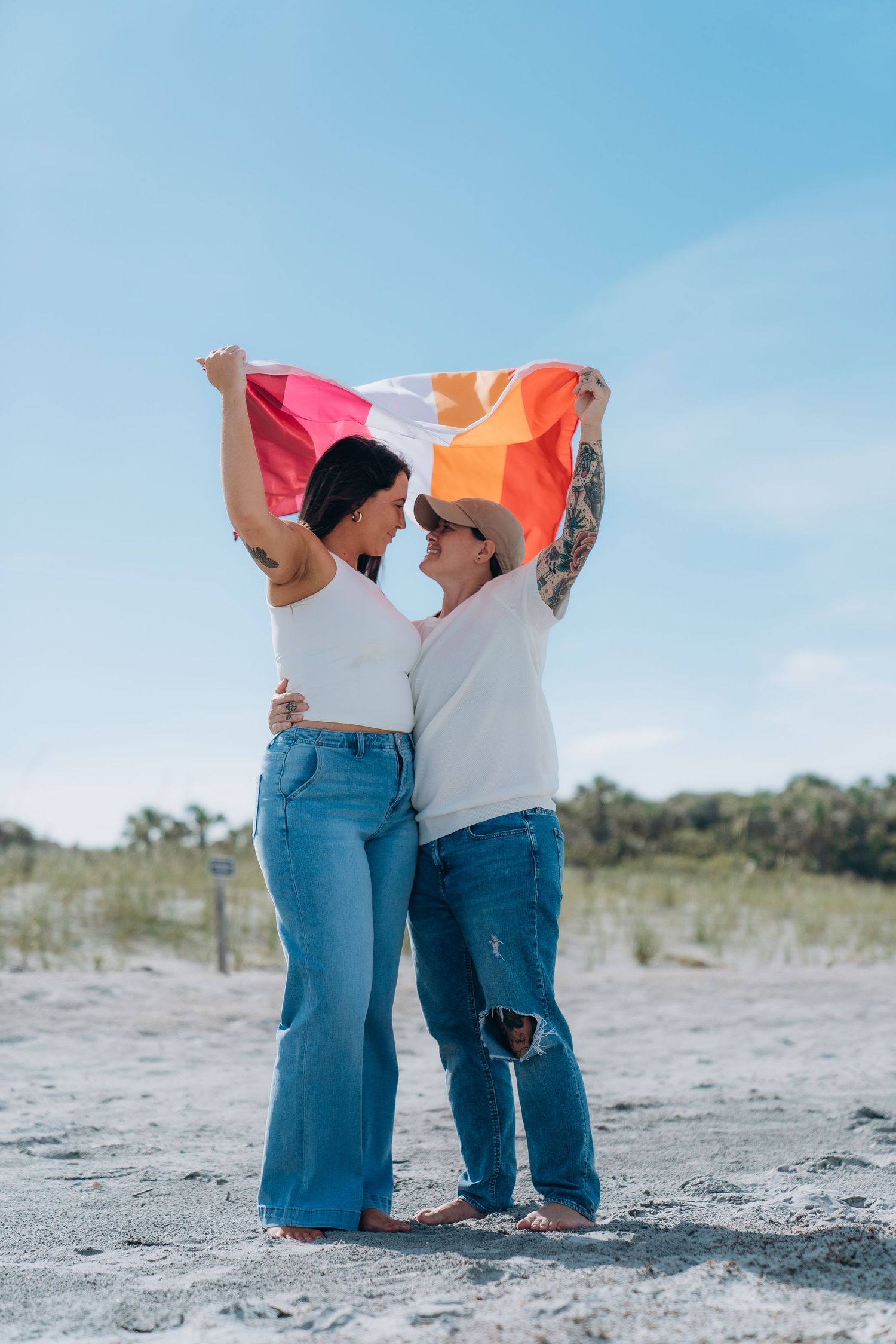 Two people in jeans and white tops standing close together on a sandy beach.