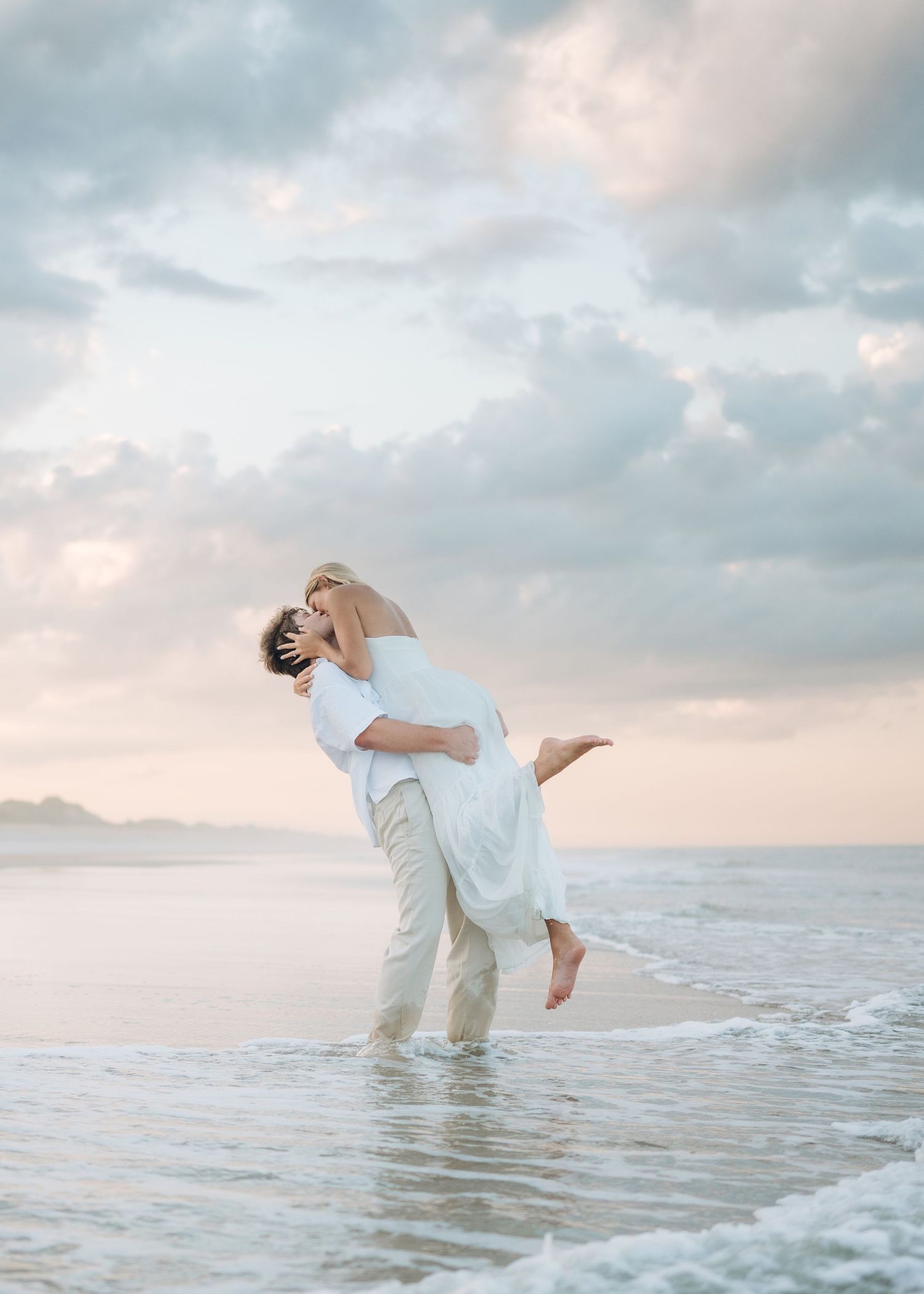 A romantic couple plays in the ocean water during sunset with cloudy skies above.