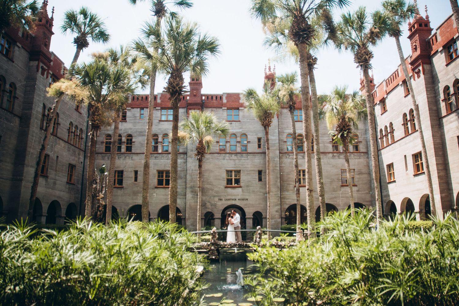 Bride and groom pose in lush courtyard of historic Flagler College castle surrounded by tall palm trees in St. Augustine, FL.