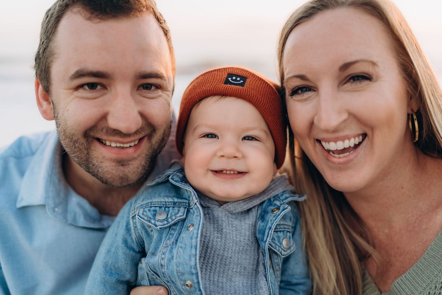 Happy family portrait with smiling parents and adorable baby wearing a rust-colored beanie outdoors.
