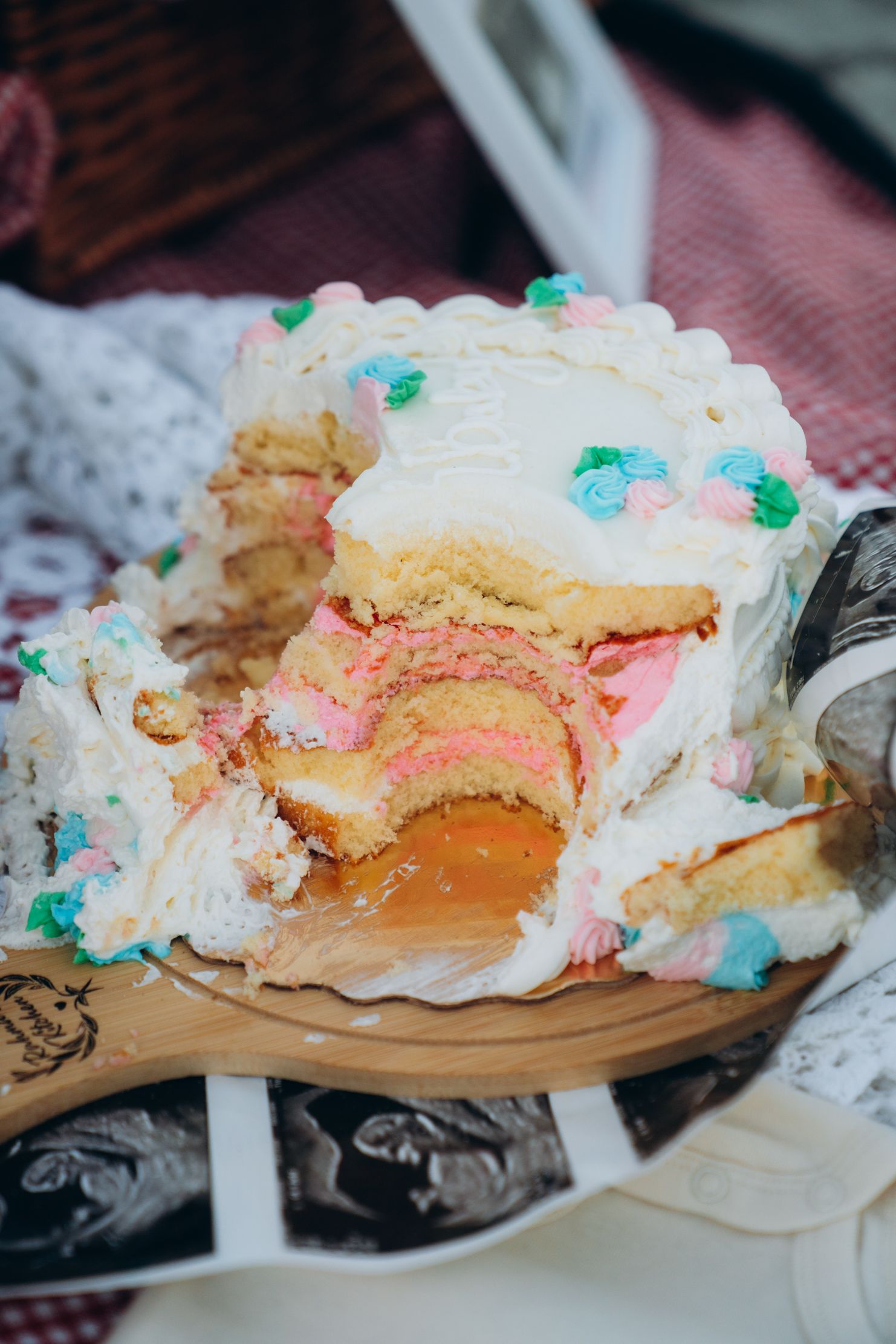 A partially sliced layered cake with white frosting, pink and white layers, and colorful flower decorations on a wooden board.