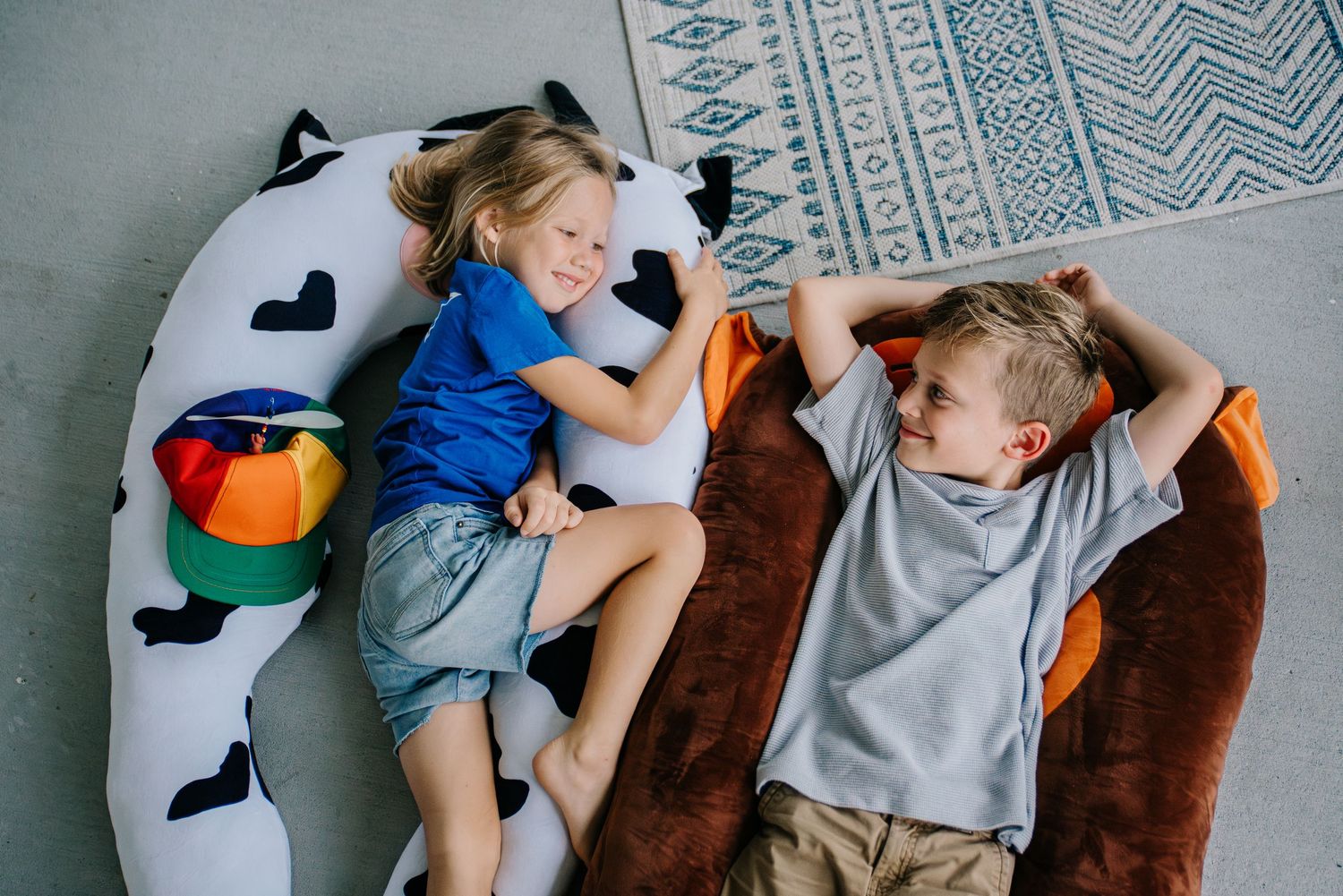 Two children relaxing on large stuffed animal pillows on a floor, smiling and playing together.