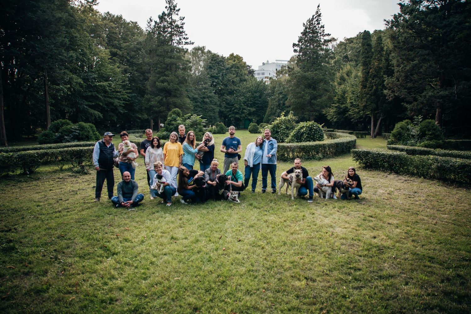 Large group of people posing together on a green lawn surrounded by lush trees and manicured hedges outdoors.