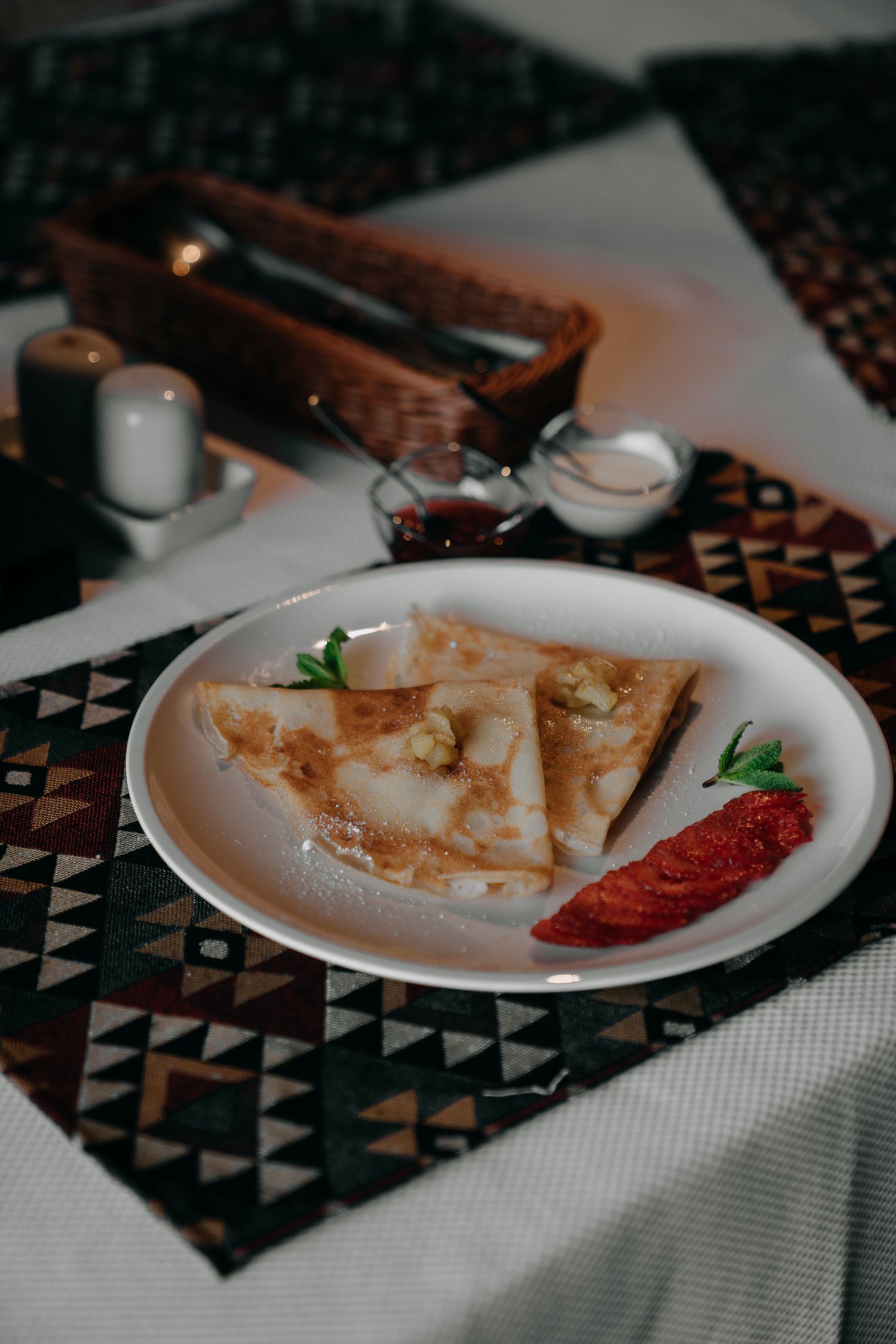 Folded crepes on a white plate with tomato garnish on a patterned tablecloth in a restaurant.