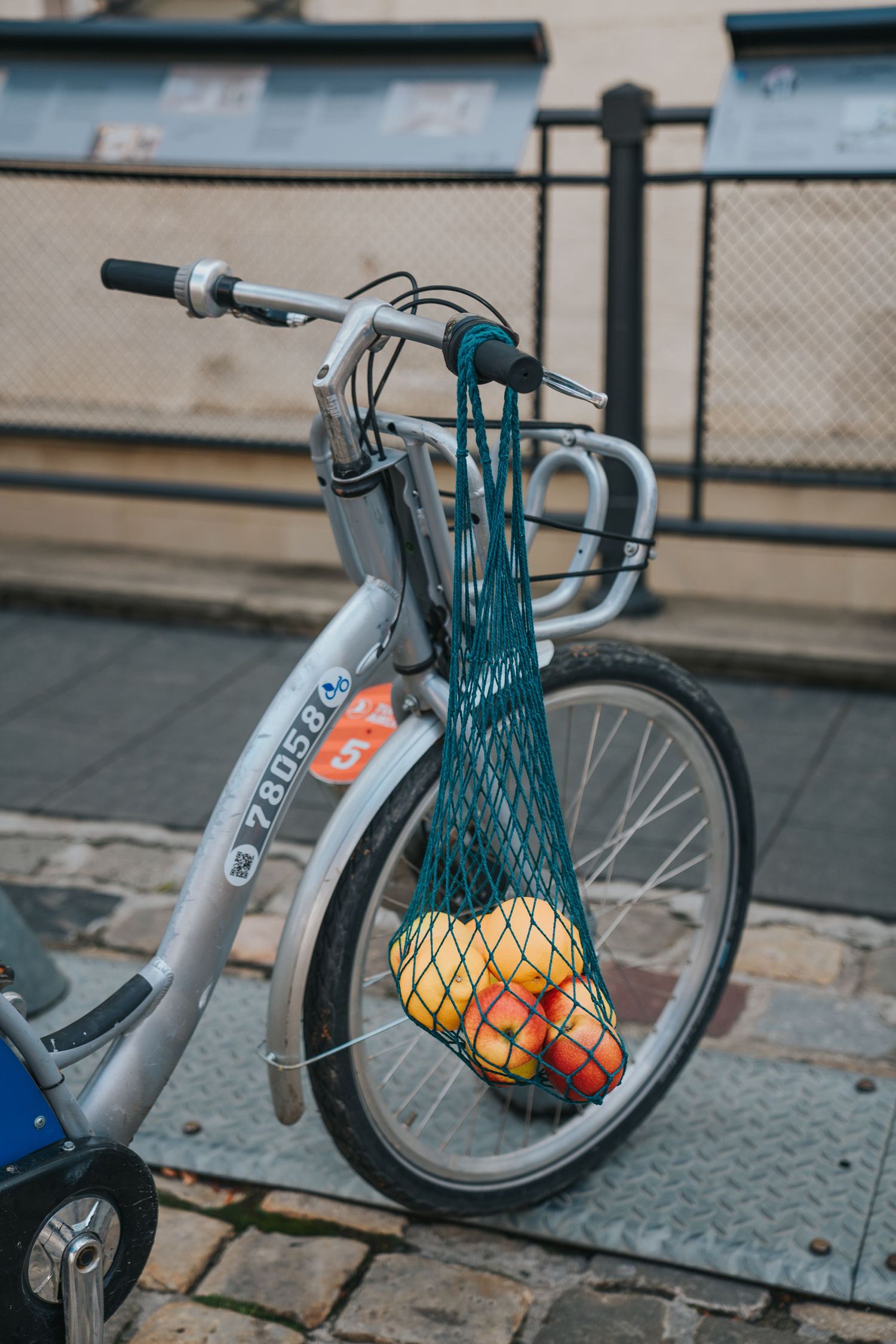 A teal mesh bag filled with colorful fruits hanging from the handlebars of a silver city bike parked on a cobblestone street.