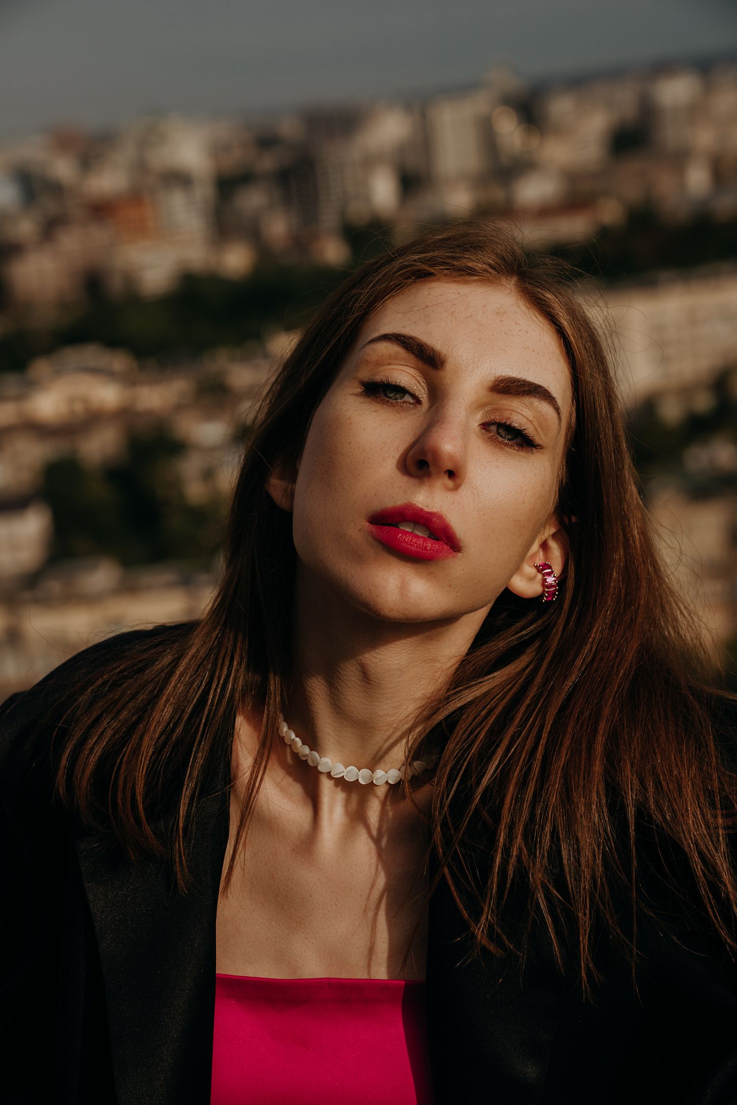Stylish woman in pink top and black jacket poses outdoors with urban hillside background.