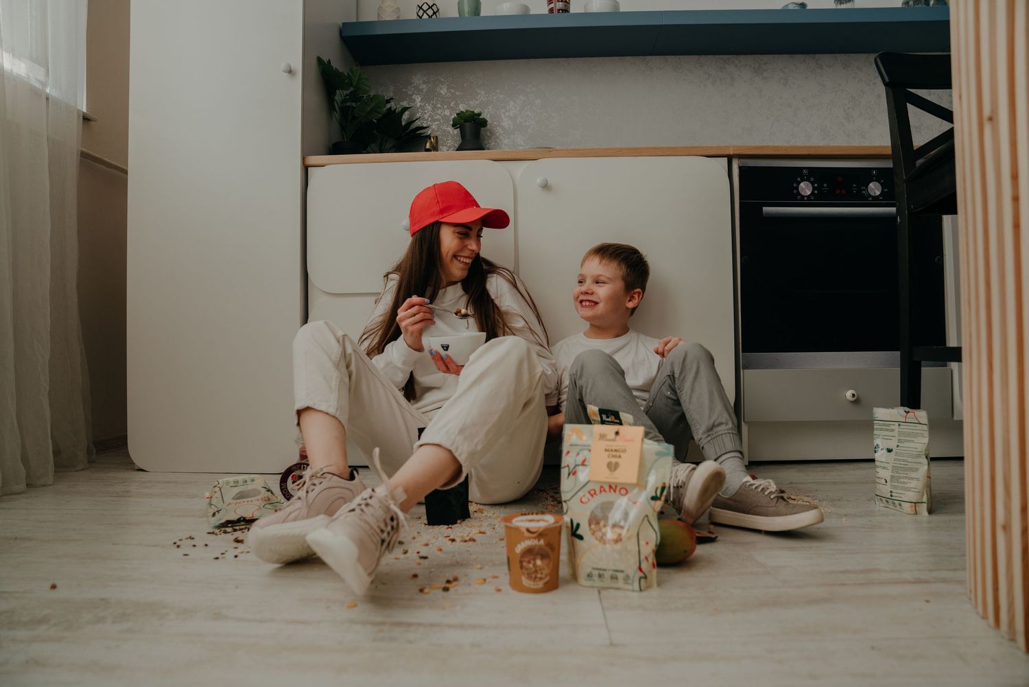 Two kids sitting on kitchen floor surrounded by snacks and coffee cups, smiling and enjoying a fun moment together.