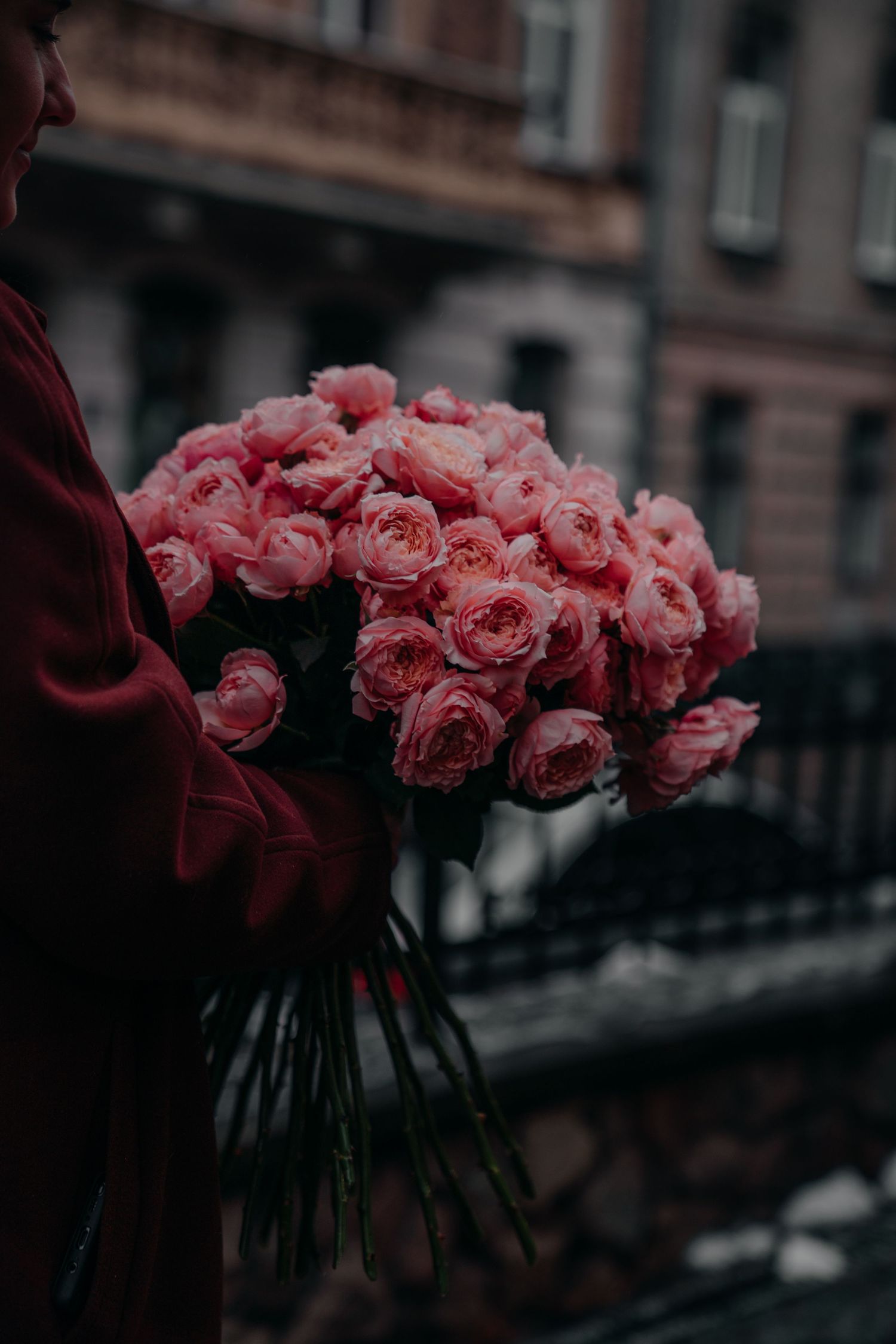 Person in red coat holding large bouquet of pink roses outdoors with blurred urban building background.