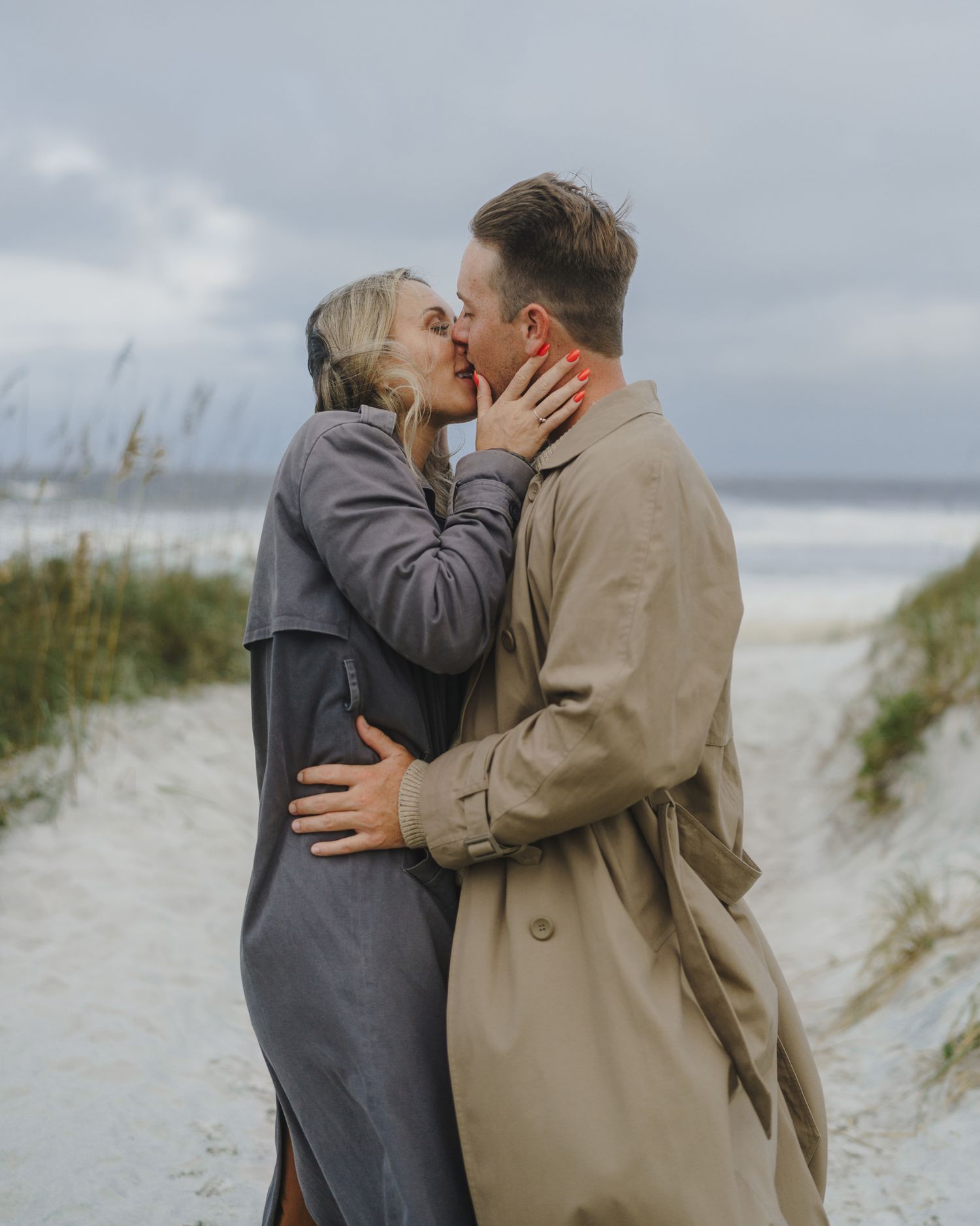 A romantic moment shared between a couple embracing on a cloudy beach with sand dunes and sea grass in the background.