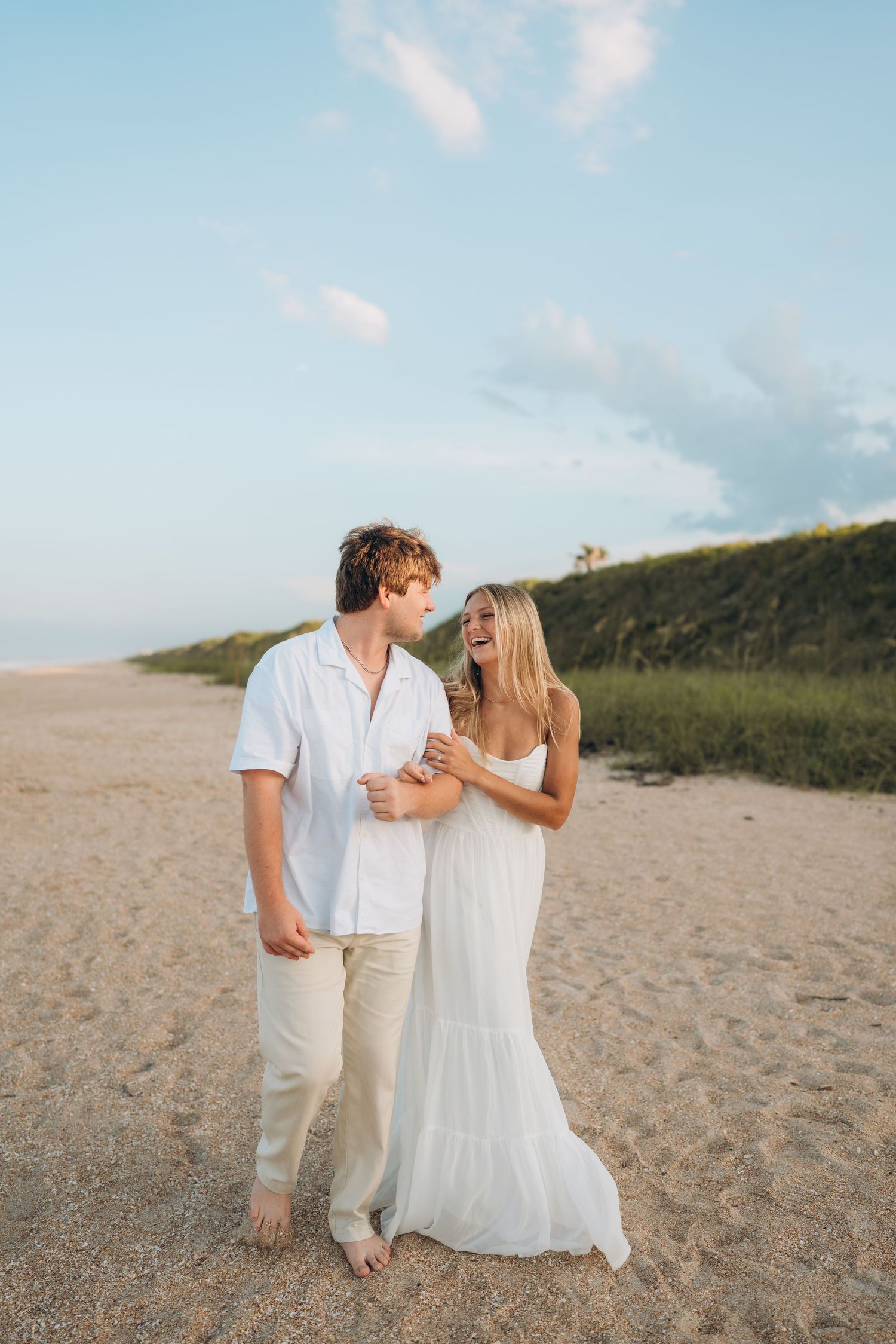 A couple in white clothing walks together on a sandy beach with hills in the background.