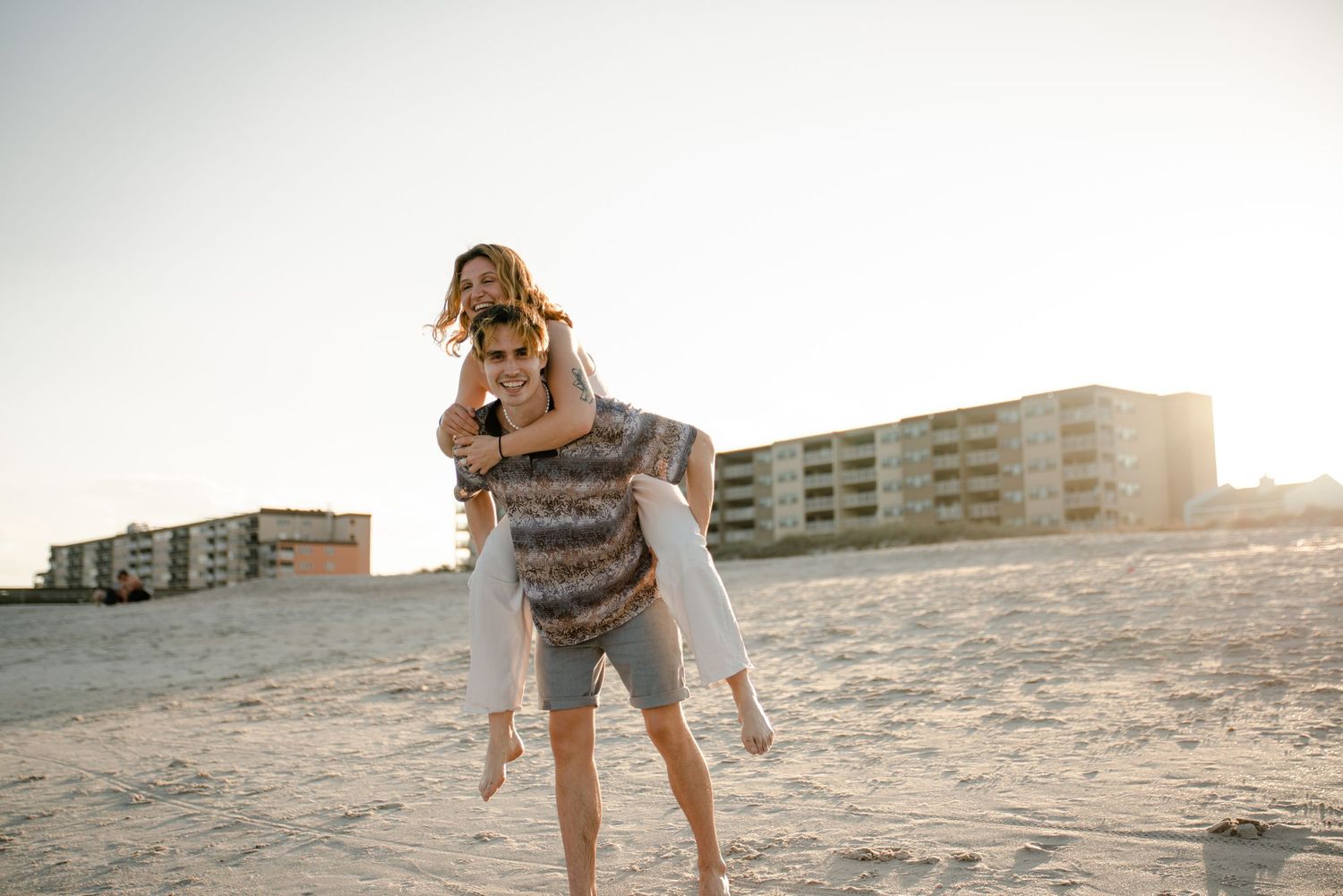 Happy couple enjoying a playful moment on beach with coastal buildings in background.