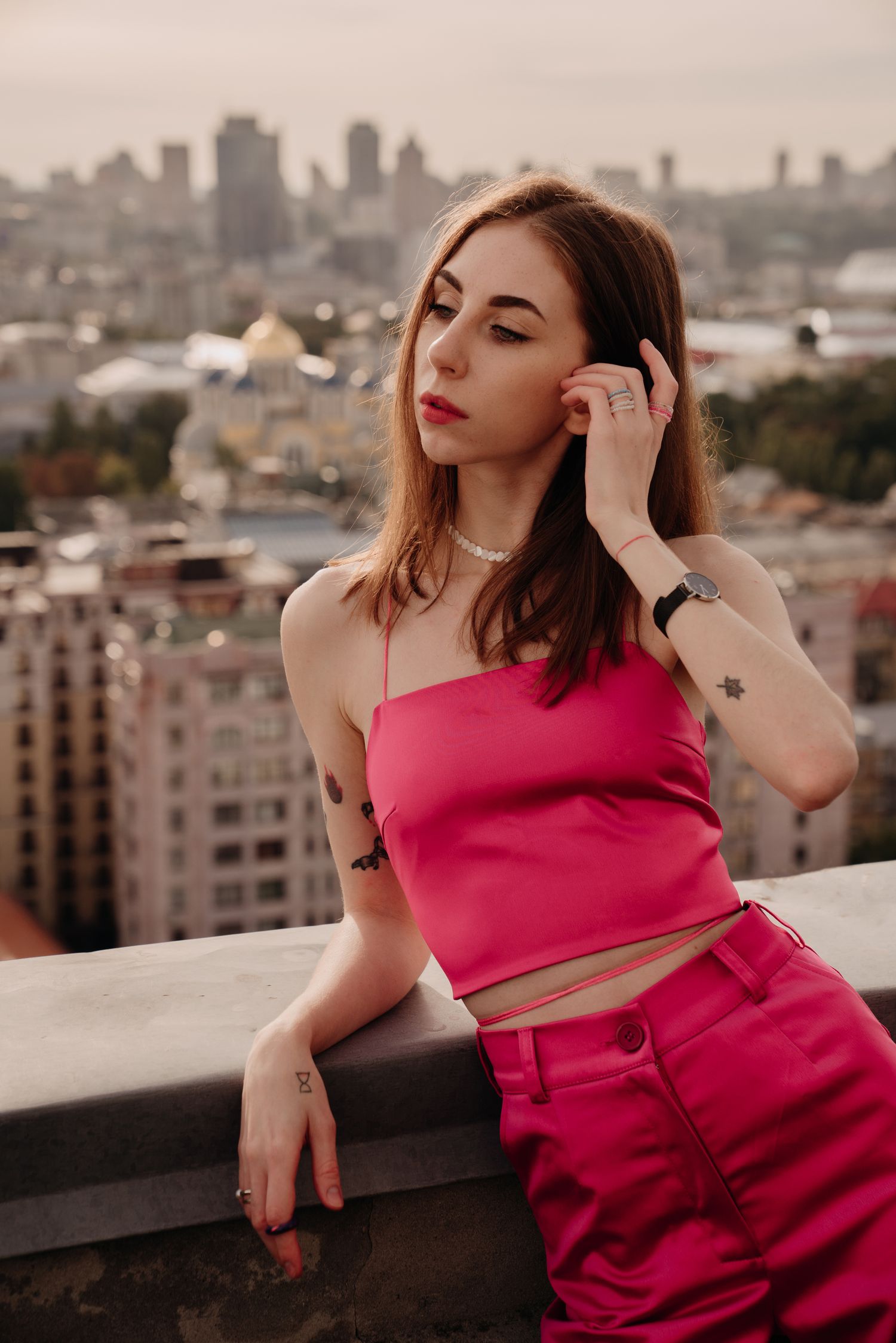 Young woman in pink crop top and pants poses on rooftop with city skyline backdrop.