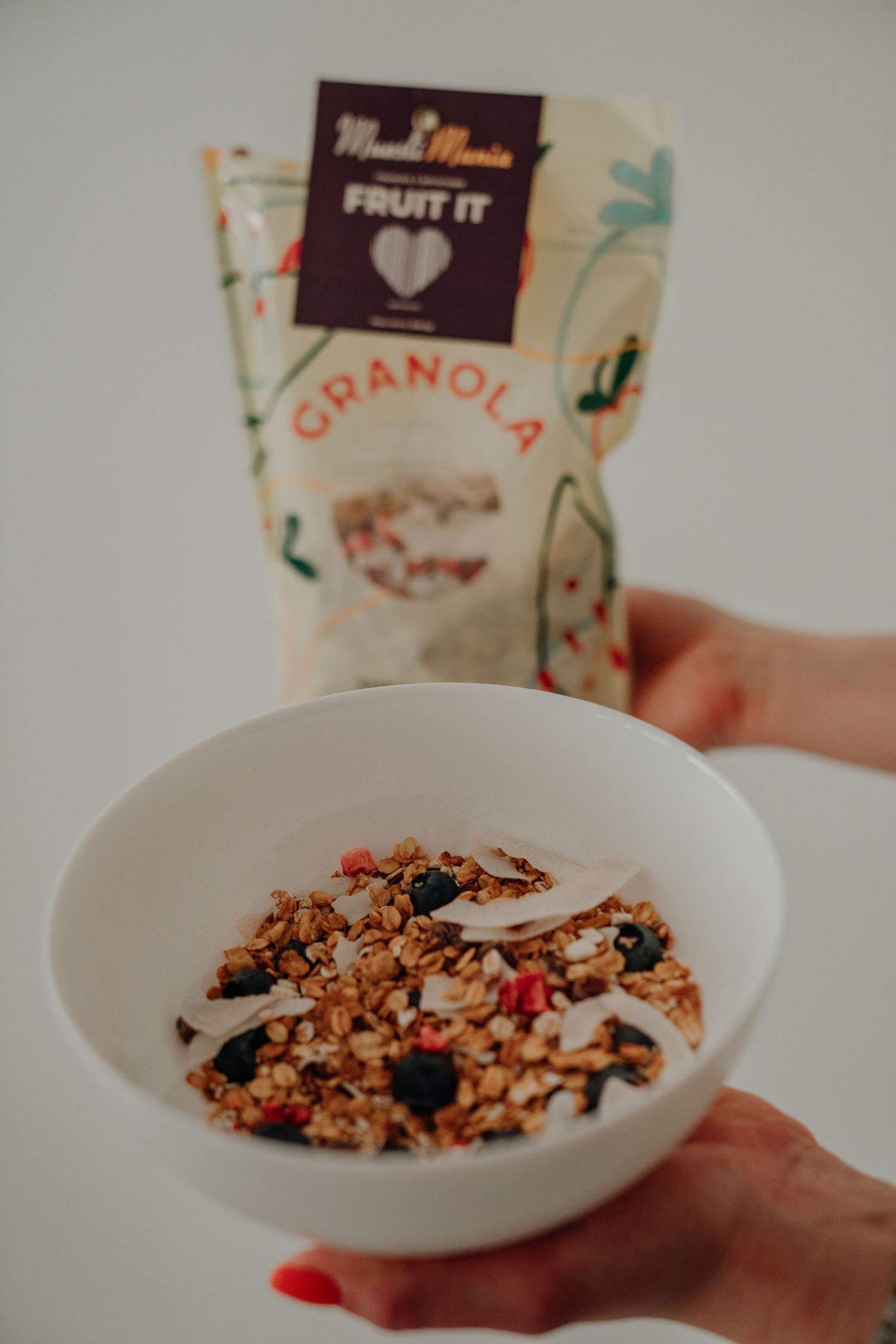 Hand holding a white bowl of Muesli Mania Fruit It granola with berries and coconut flakes, with bag in background.