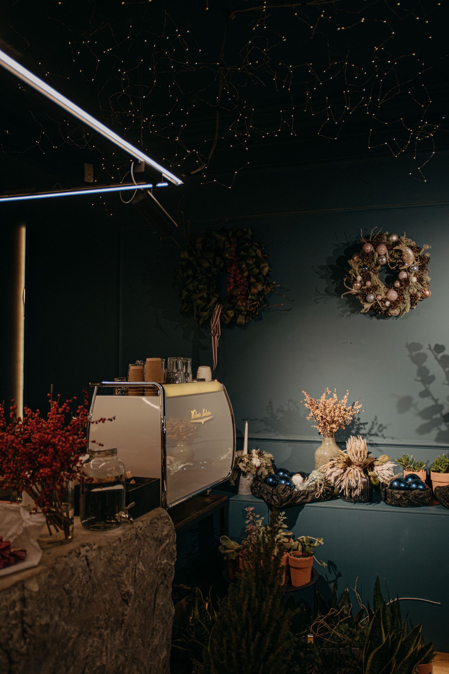 Cozy dark cafe corner with espresso machine, dried flower wreaths on teal wall, and starry ceiling lights.