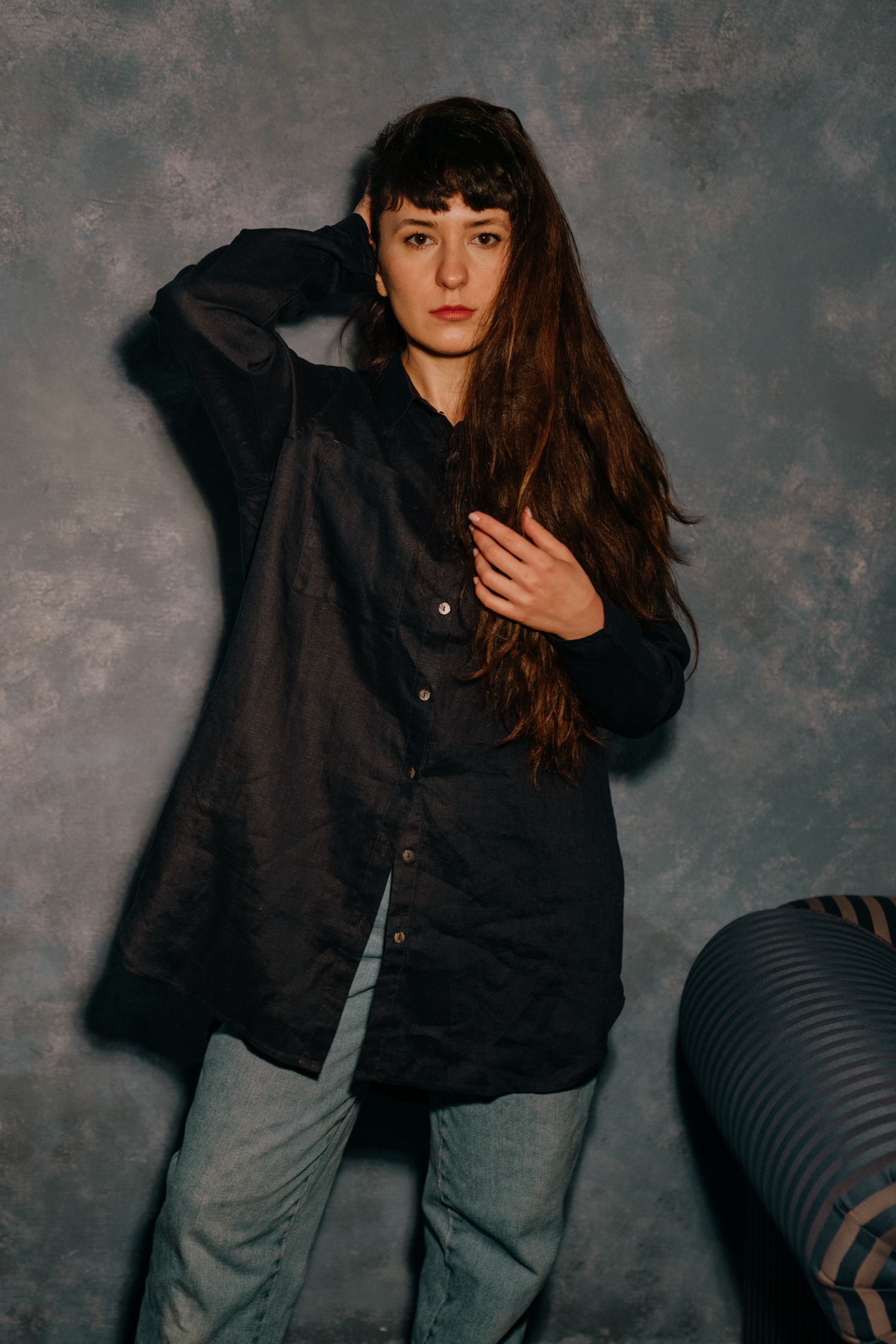 Woman in oversized black button-up shirt and light jeans posing against gray textured studio backdrop.