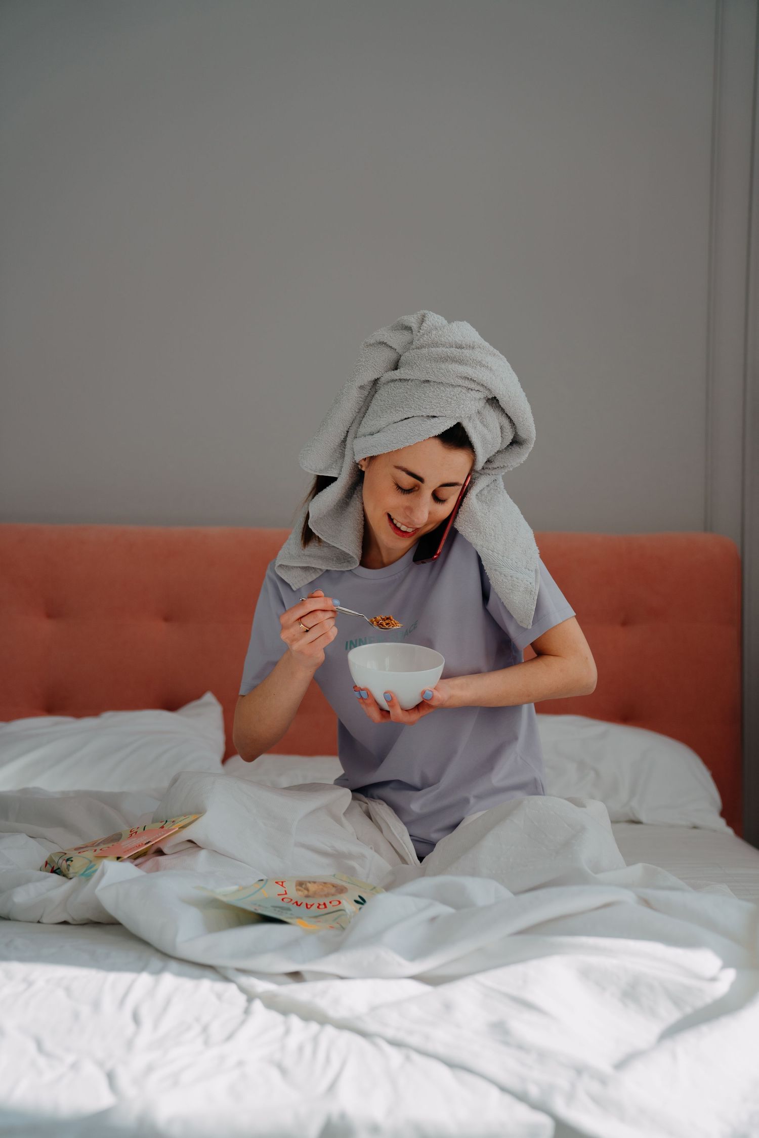 Woman with towel on head sitting in bed eating granola from a white bowl.