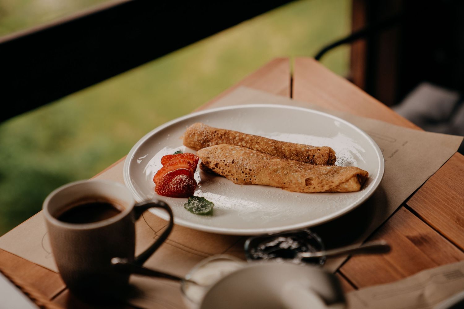 A white plate with two crepes, fresh strawberries, and herbs beside a coffee cup on a wooden table.