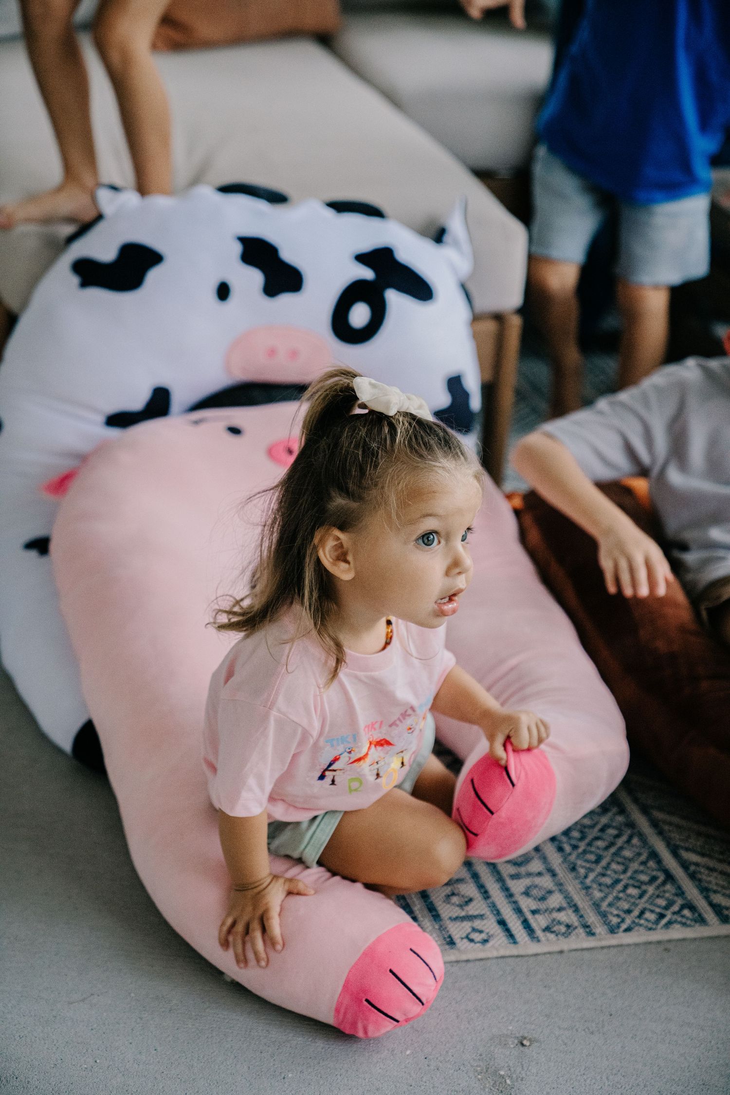 Toddler girl sitting on large pink pig plush pillow on floor indoors.