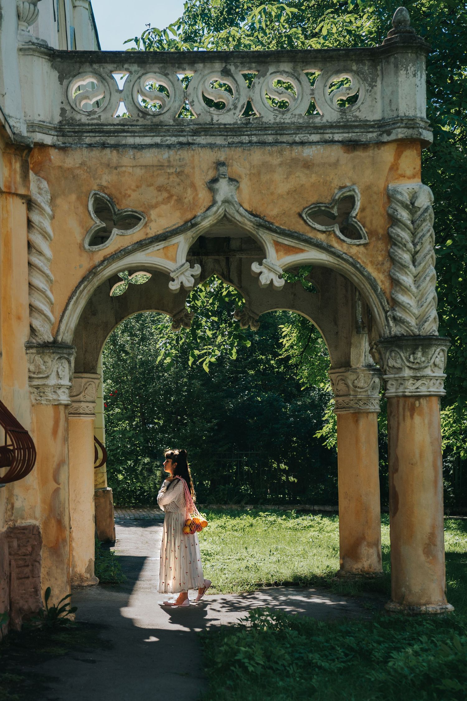 Woman in floral dress standing under ornate stone arch gateway surrounded by lush green trees in sunlight.