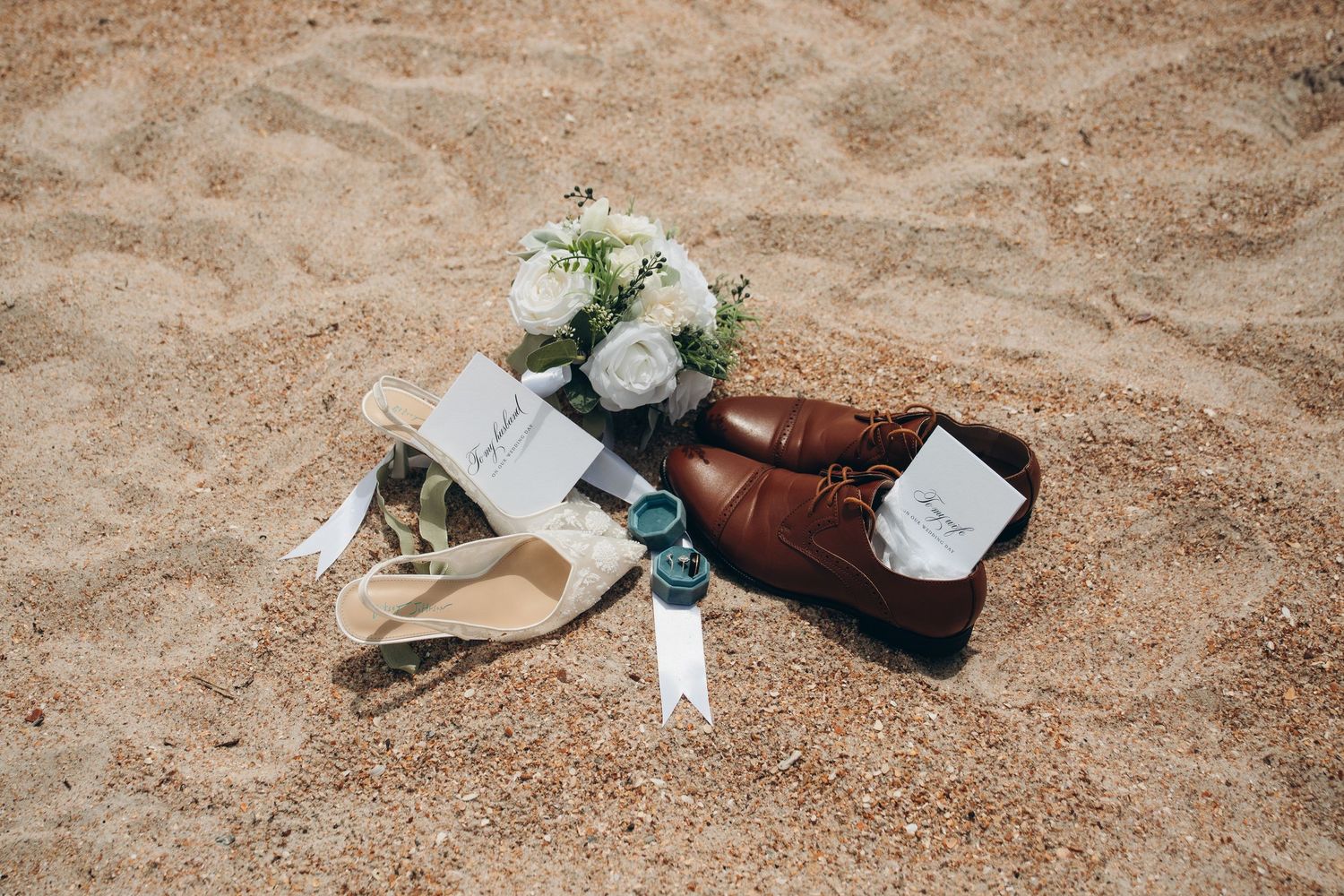 Wedding flat lay on sandy beach featuring brown leather dress shoes, white floral bouquet, place cards, and accessories.