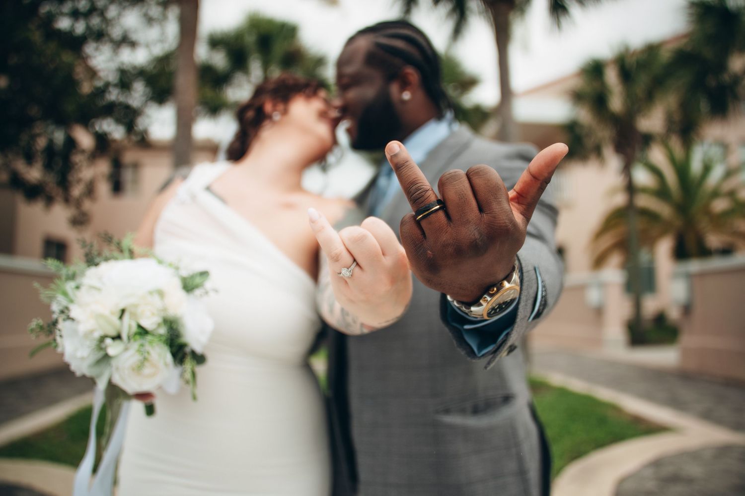 Newlywed interracial couple showing wedding rings, bride holding white bouquet, groom in gray suit, outdoor palm tree setting.