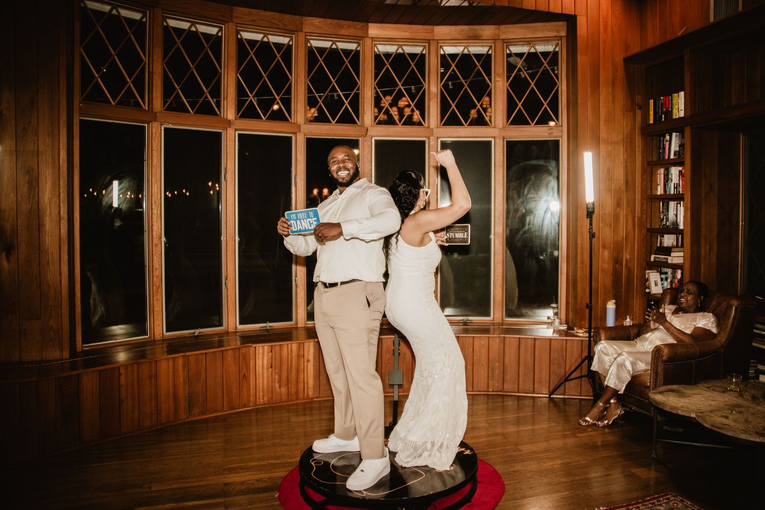 Couple dancing joyfully on a small stage in a warmly lit wood-paneled room during their wedding reception.