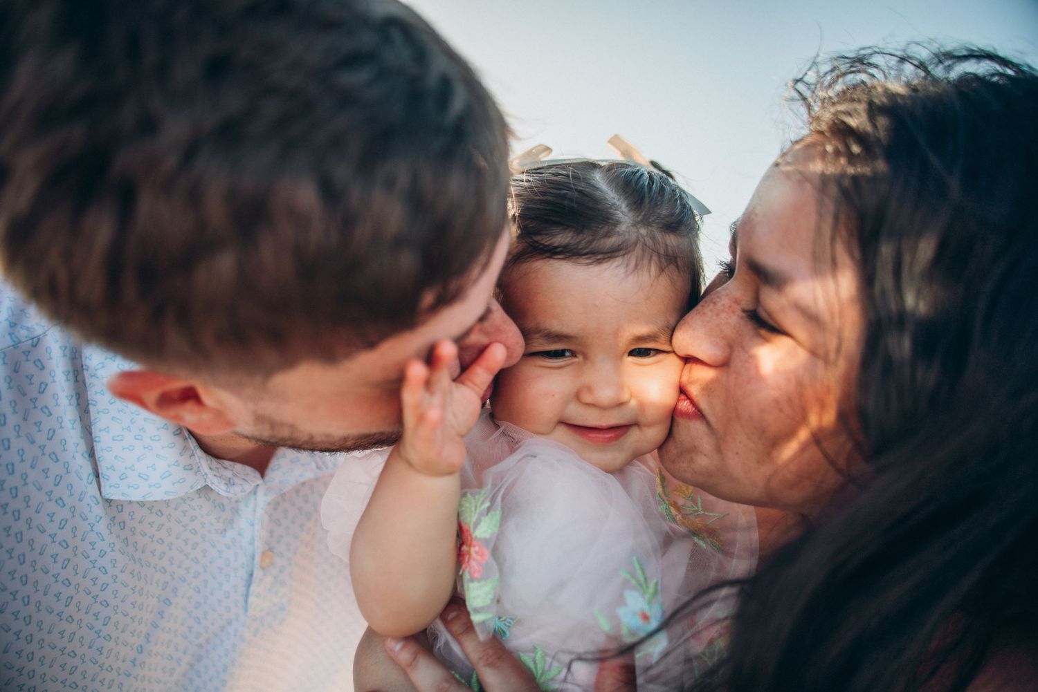 Parents kissing their smiling baby girl on both cheeks in a heartwarming family portrait outdoors.