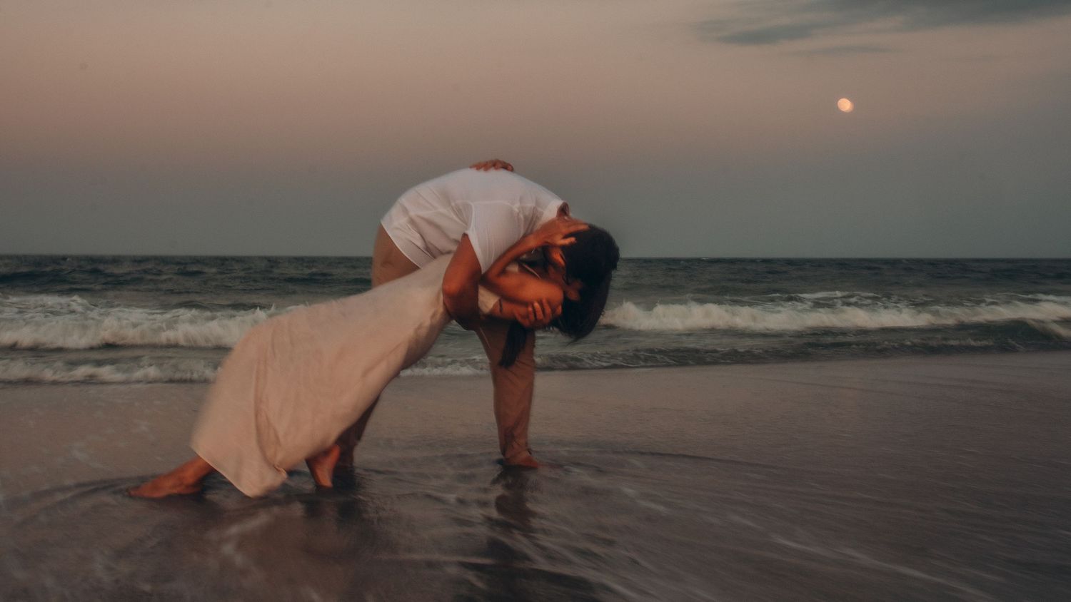 Romantic couple sharing a passionate kiss on a beach at sunset with ocean waves and a glowing moon in the background.