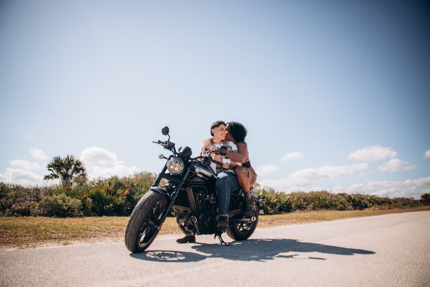 Couple kissing on a classic black motorcycle parked on a sunny road with blue skies and green trees.