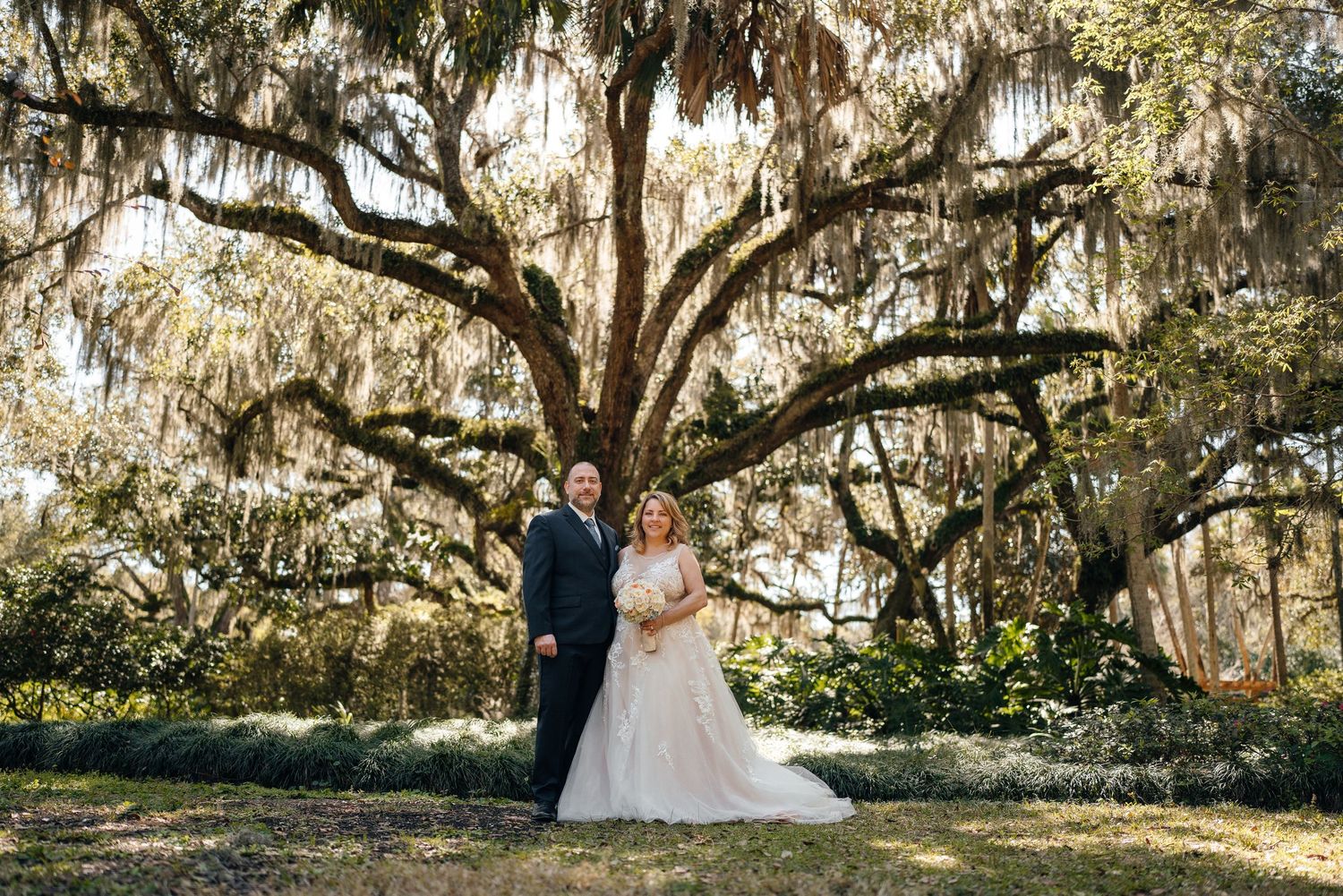 Bride and groom pose under a grand moss-draped oak tree in a lush garden setting.
