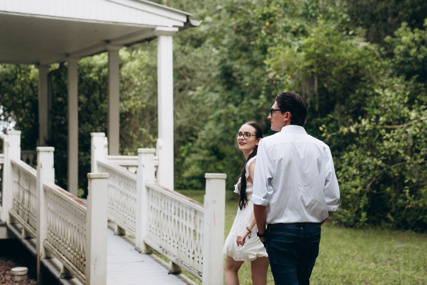 A couple embraces on a white porch with columns and railing surrounded by green trees.